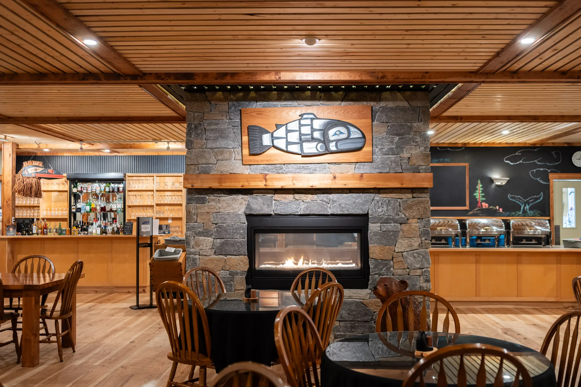 The lounge at Knight Inlet Lodge from the far side, the stone fireplace lit and the dining tables beyond