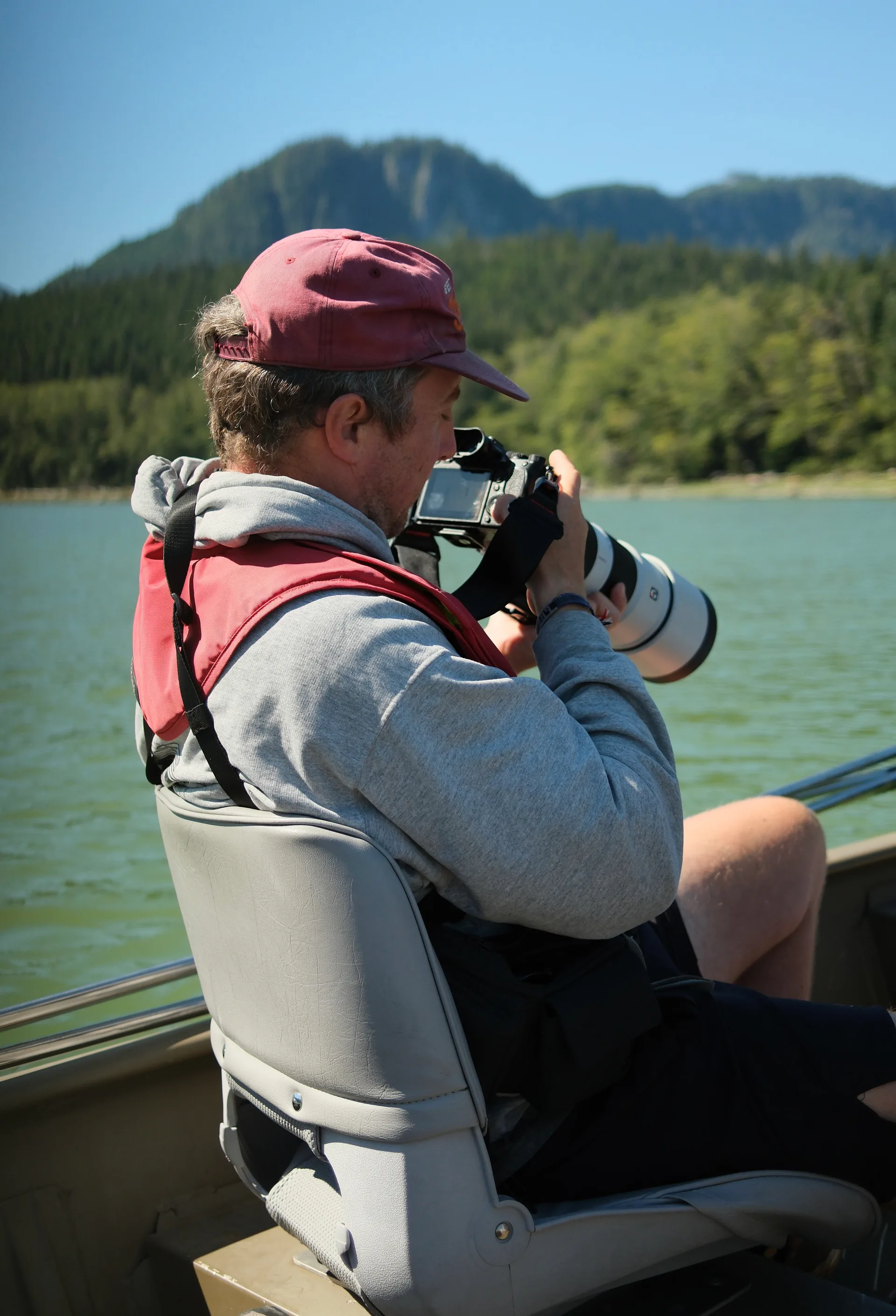 The author seated in a skiff in a red cap and life jacket, the Sony A1 and white 200-600 raised to his eye, forested hills across green water