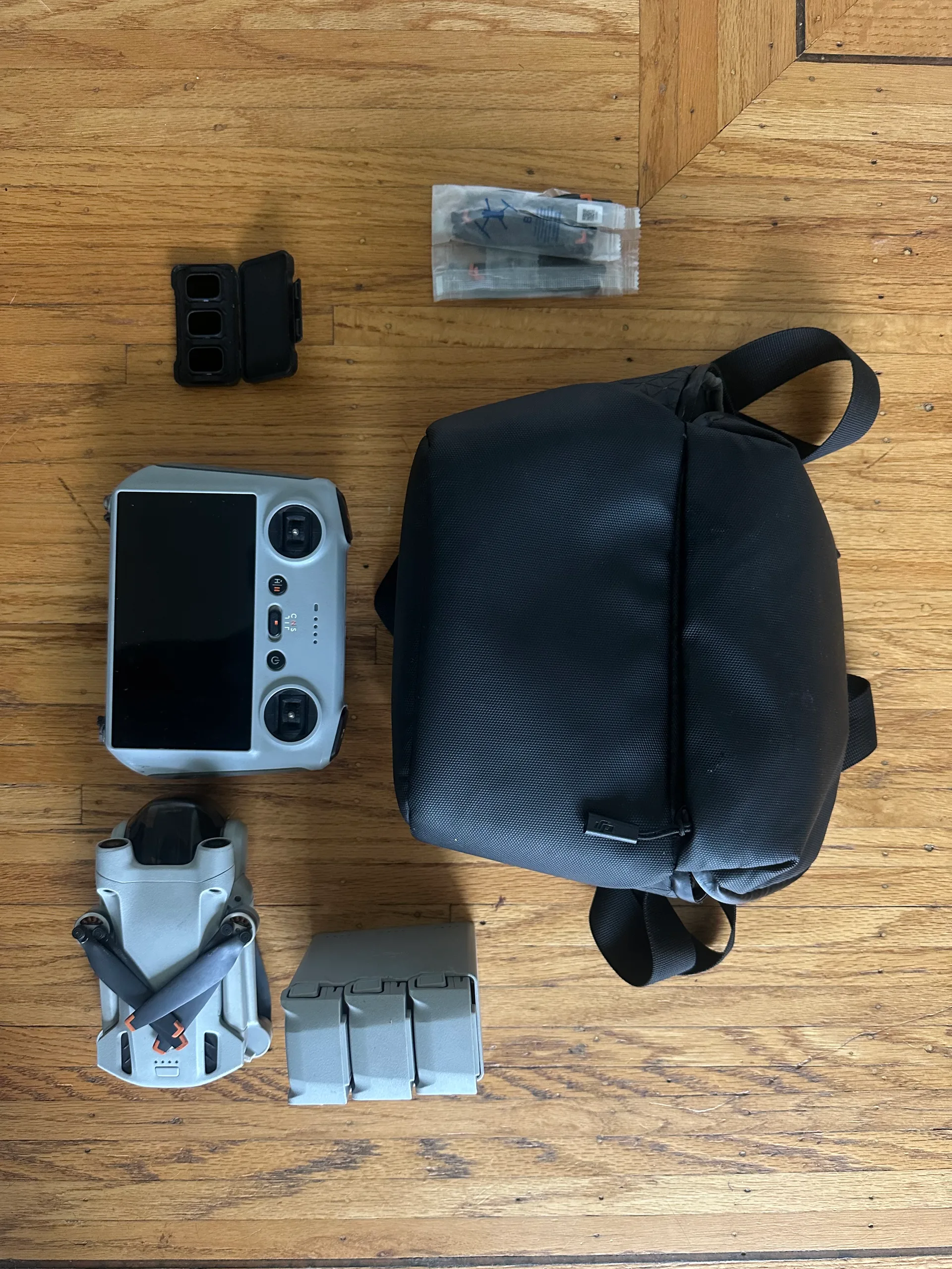 DJI Mini 3 Pro kit laid out on a wooden floor: the folded drone, the RC controller with screen, three batteries in their charging hub, a case of ND filters, spare propellers in a bag, and a black shoulder bag