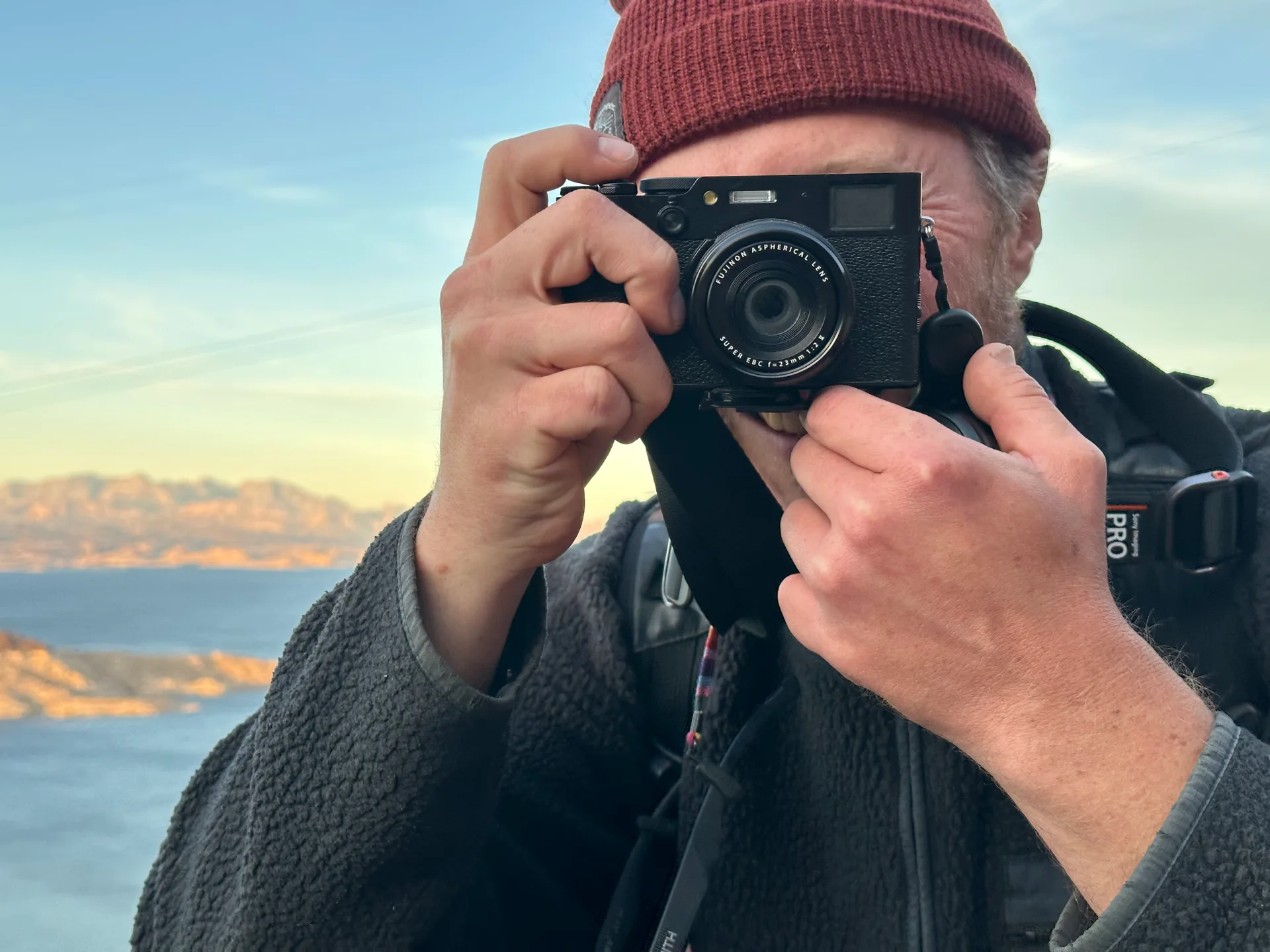 The Fujifilm X100VI held to the eye in both hands, a rust-coloured knitted hat above it, sunlit mountains and sea behind