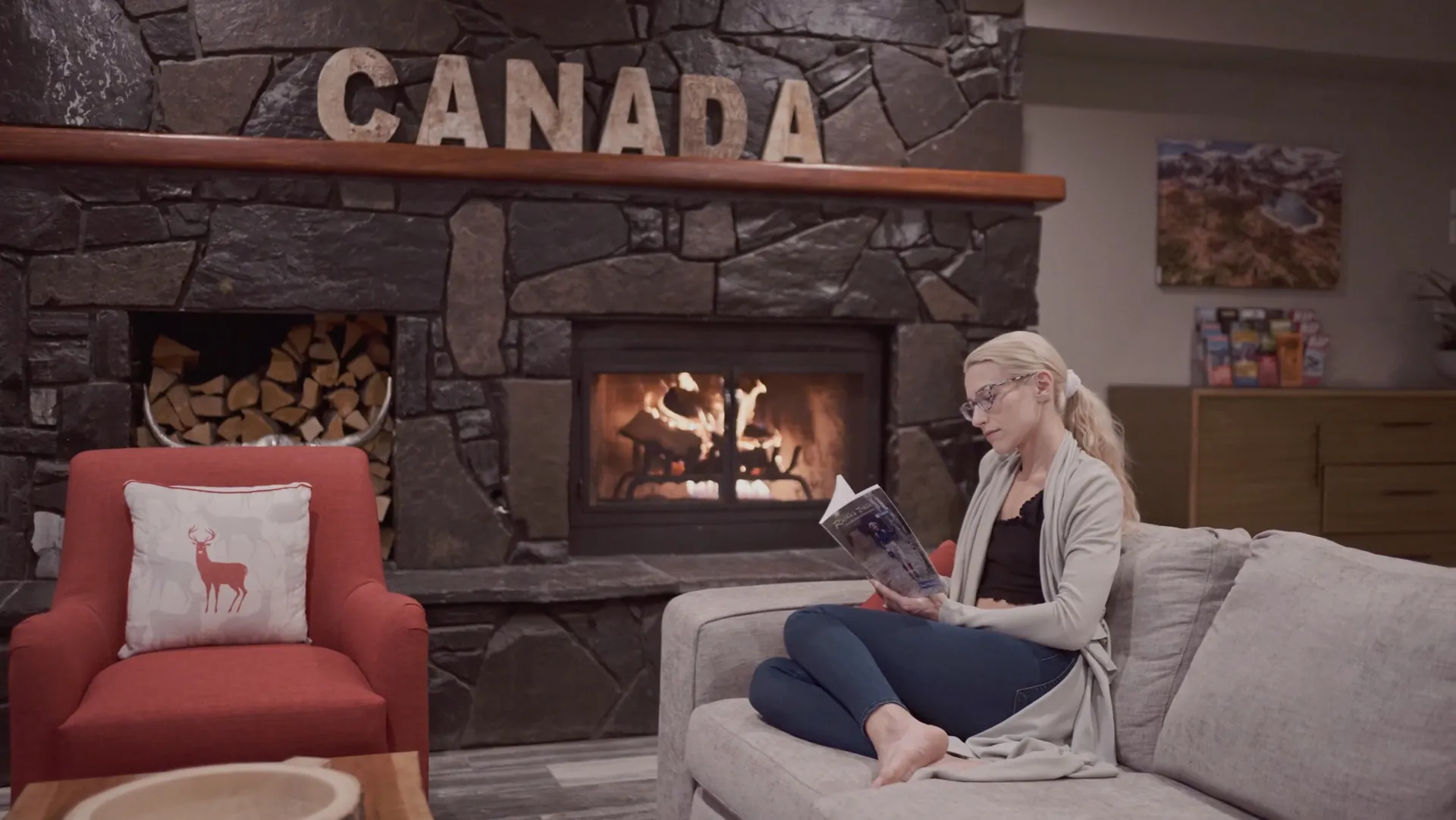 A guest reading on the sofa beside the lit stone fireplace in the great room