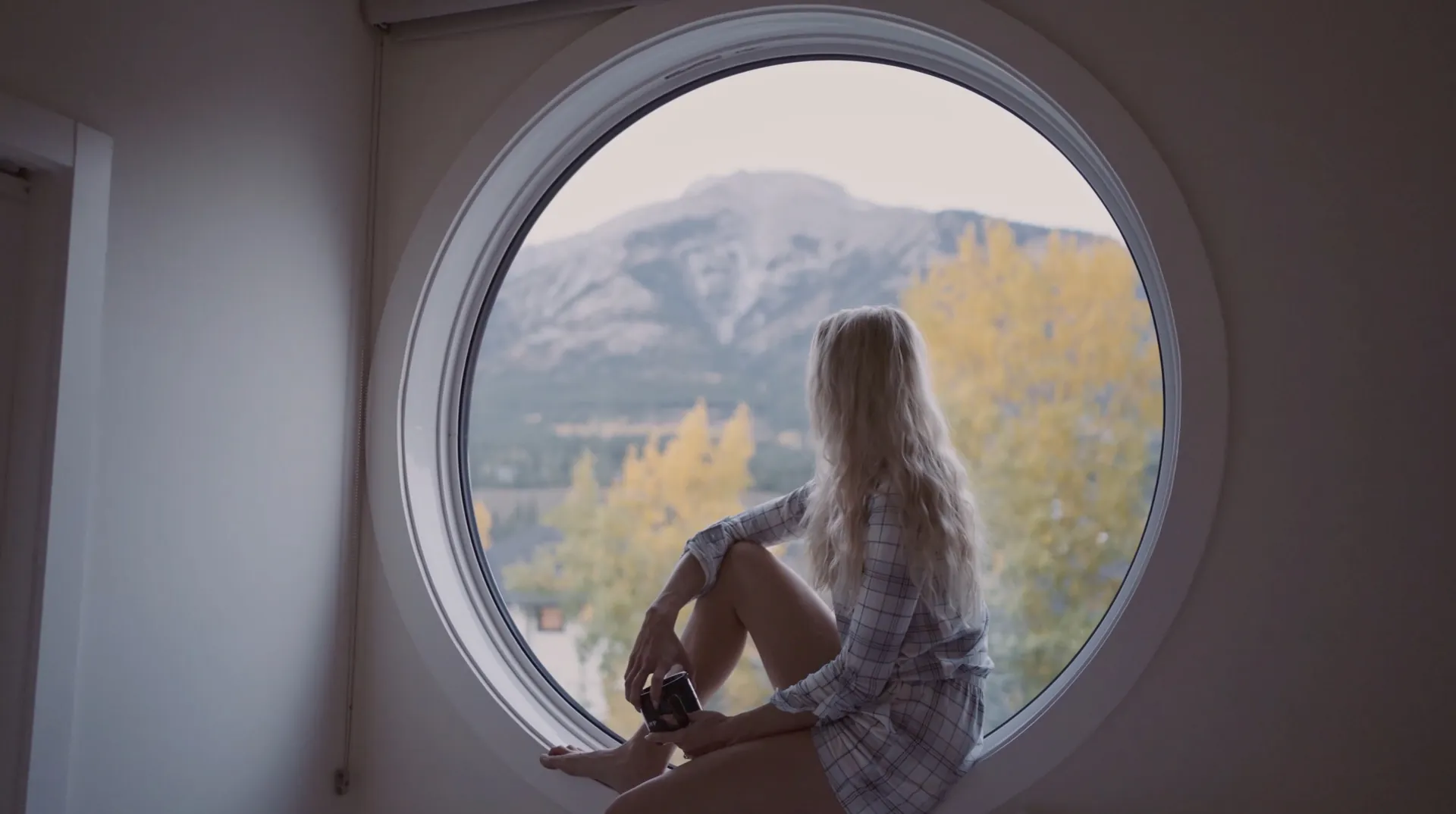 A guest sitting in the signature round window of a Basecamp Resorts Canmore suite, autumn larches and Ha Ling Peak outside