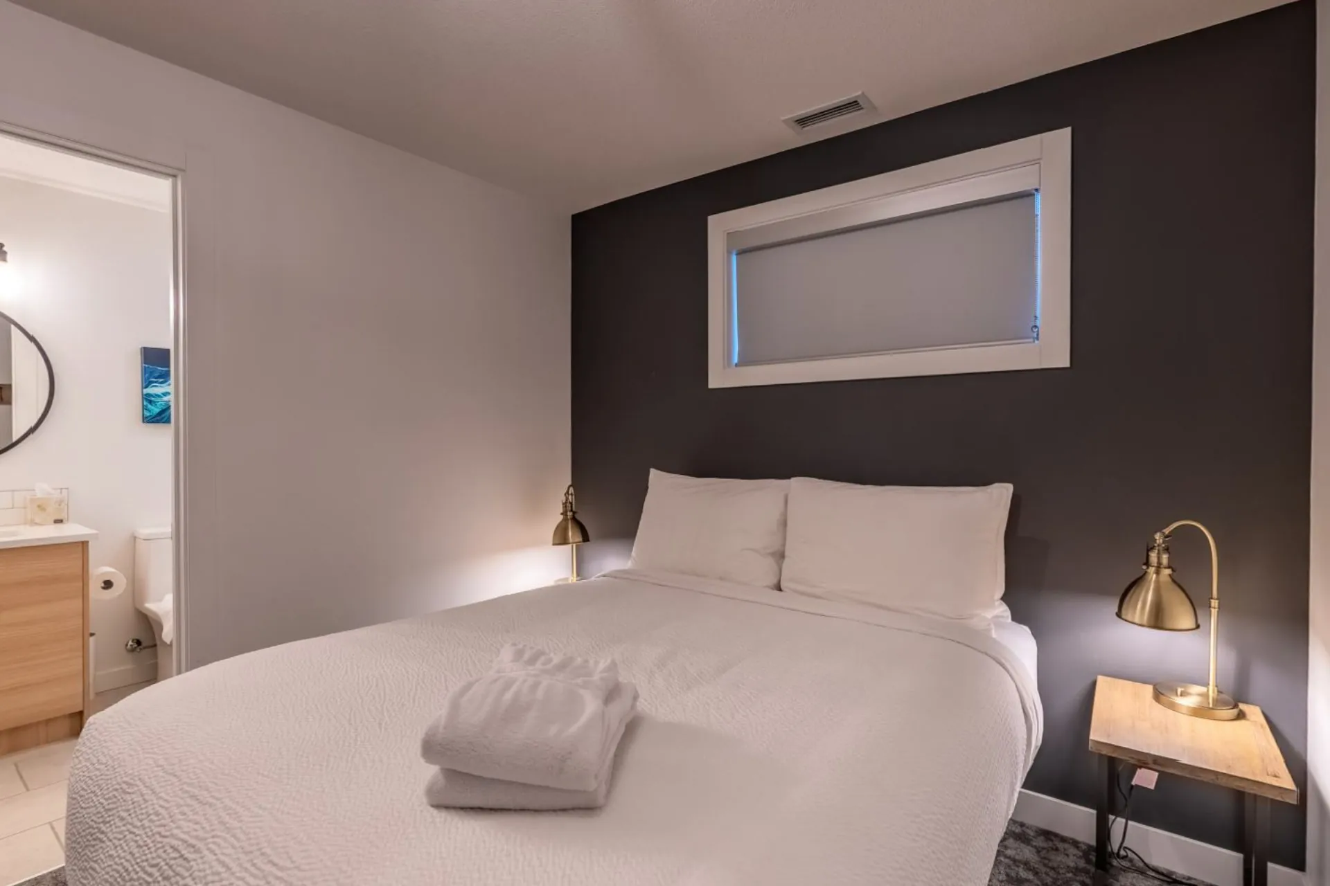 A bedroom with white linen, dark feature wall, reading lamps and a window with the forest outside