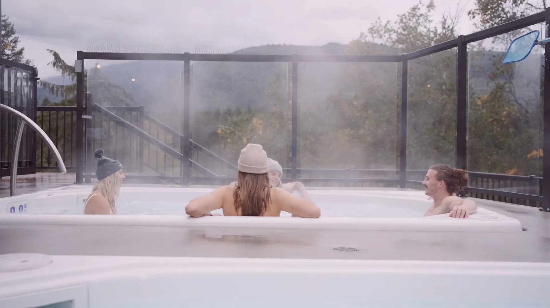 Three guests in the rooftop hot tub at Basecamp Resorts Revelstoke, steam rising, the glass rail and the mountains behind