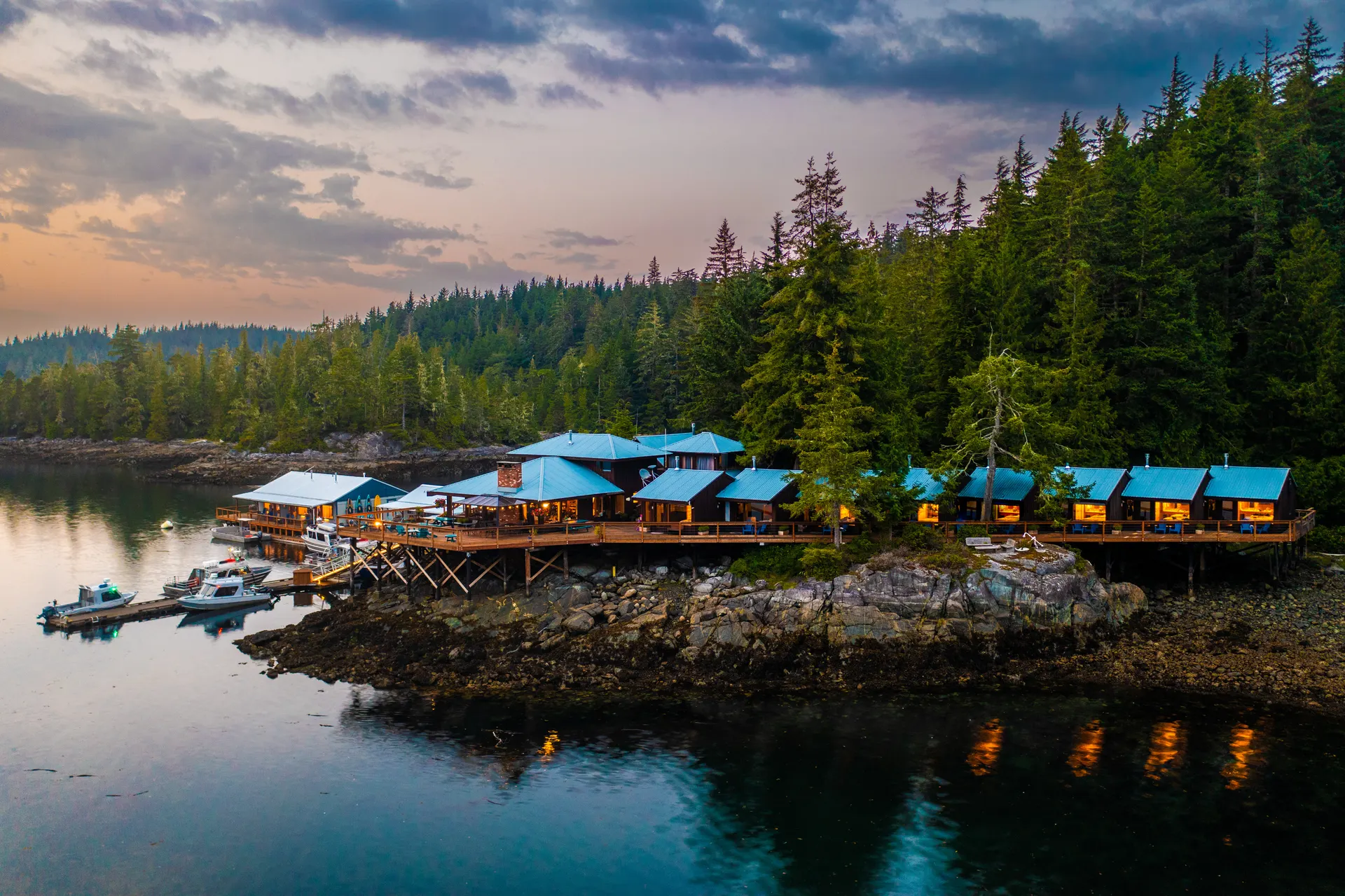 Aerial view of Berry Island Lodge at dusk, its blue-roofed cedar buildings and boardwalk of guest rooms lit along a rocky point, with boats at the dock below