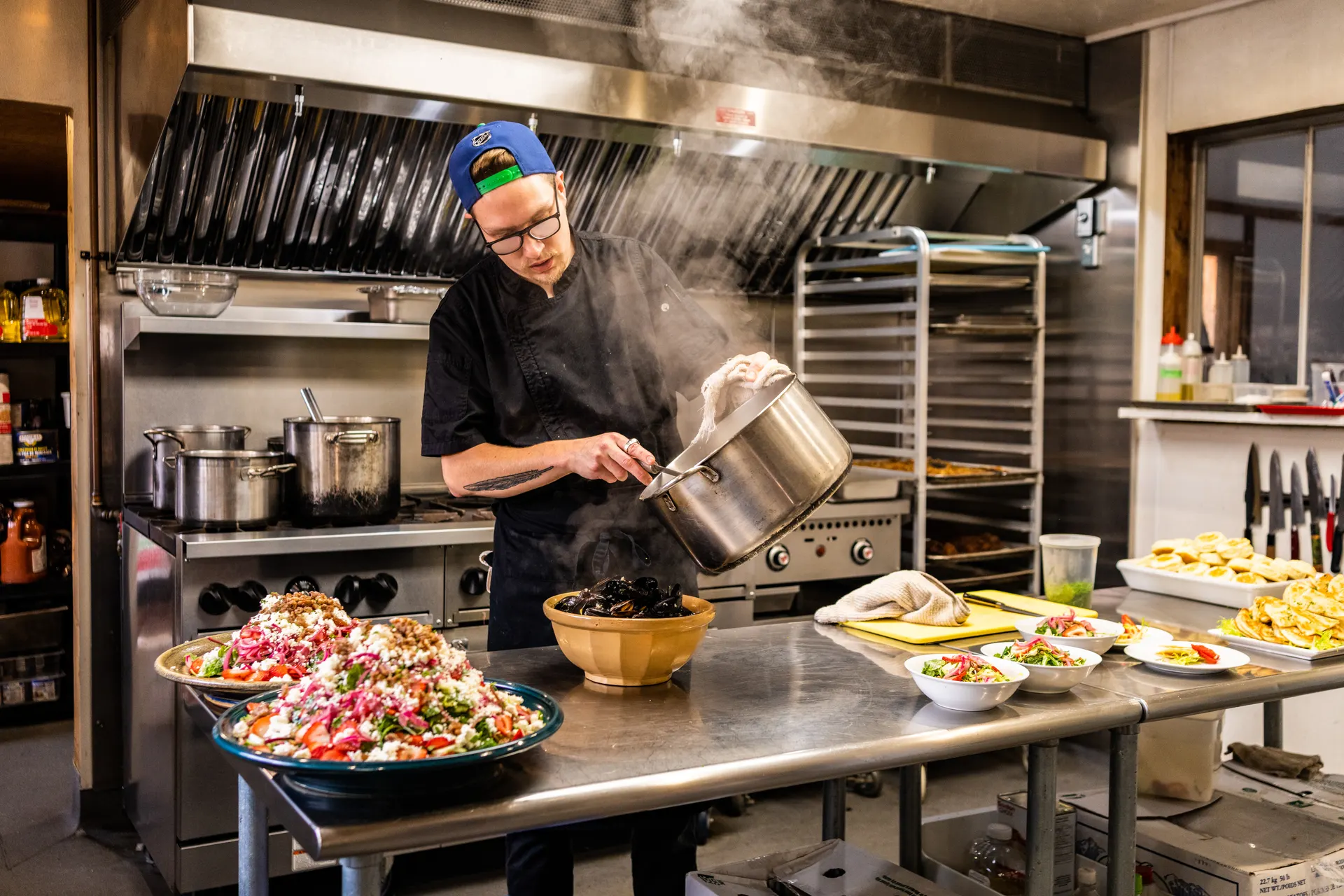 The chef pouring steamed mussels into a bowl in the lodge kitchen, a large tomato salad beside the stove