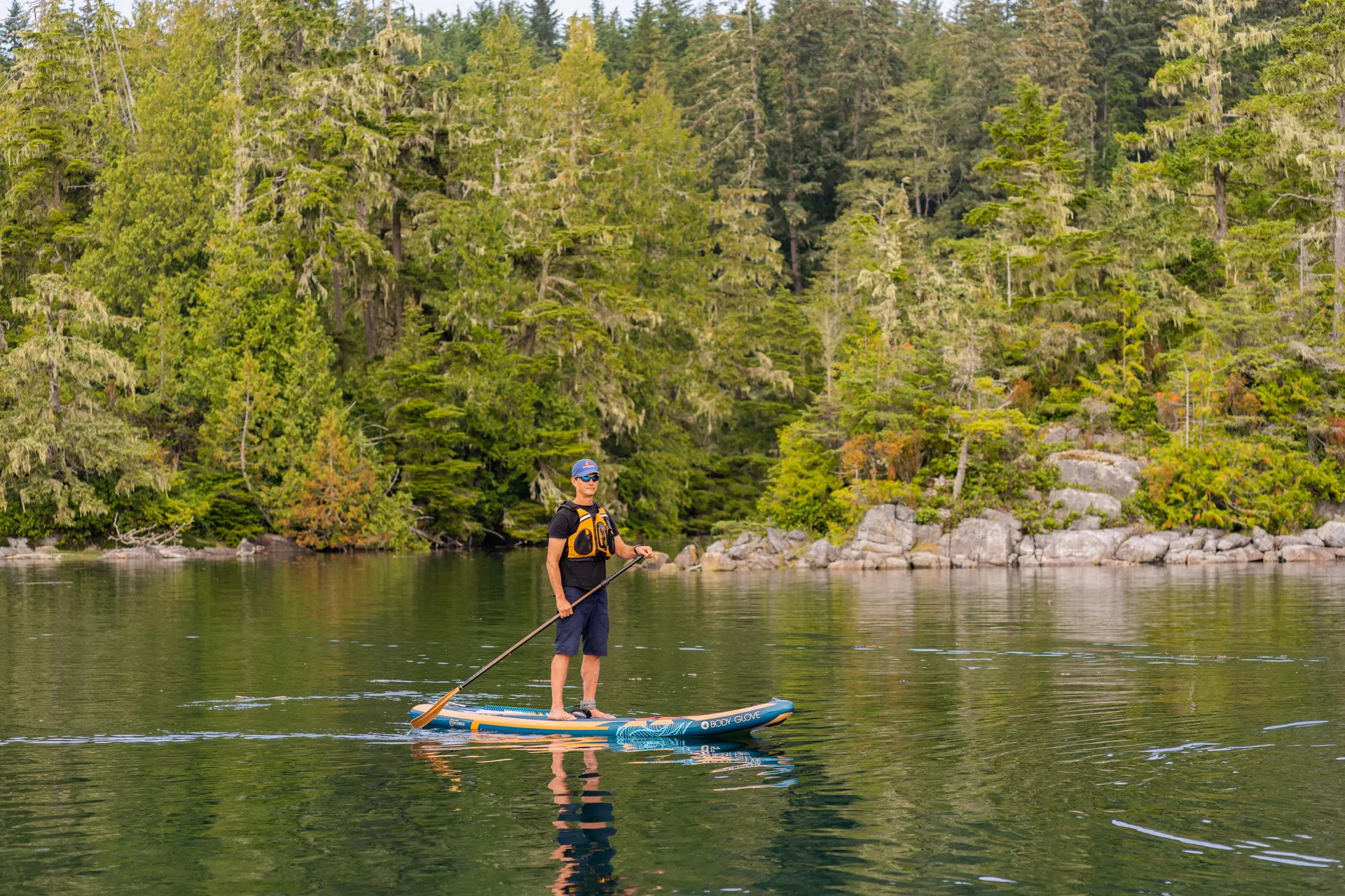 A guest on a stand-up paddleboard on calm water below a wall of old-growth forest