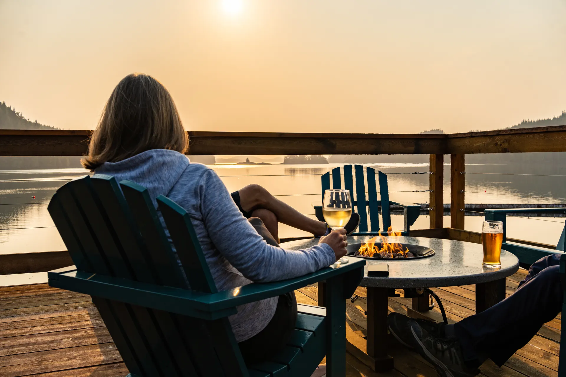 A guest in an Adirondack chair with a glass of wine on the deck at sunset, a fire table burning beside her