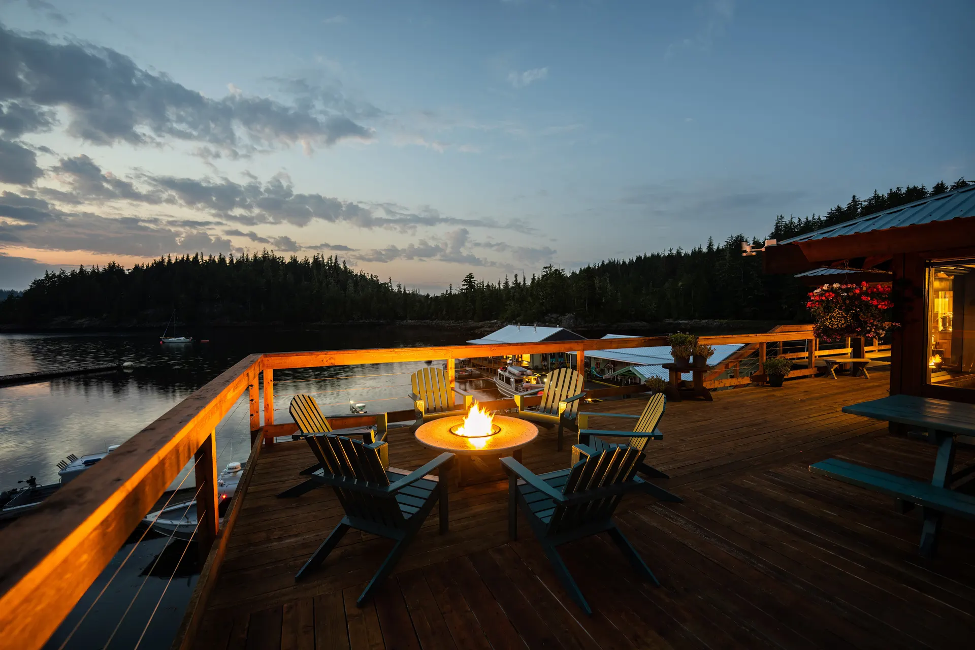 The lodge's cedar deck at dusk with Adirondack chairs around a lit fire table, looking out over the harbour