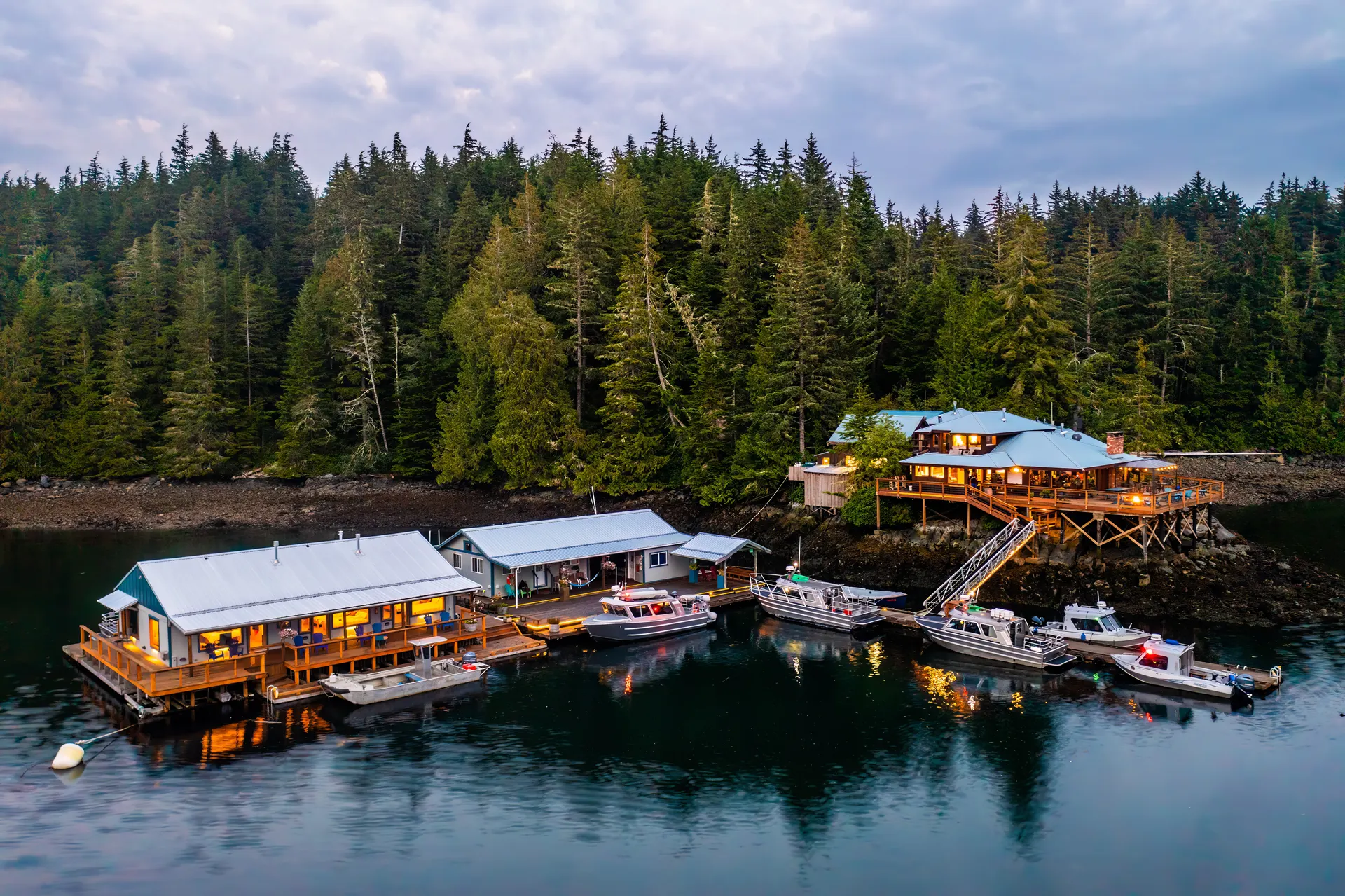 Aerial view of the floating lodge and dock at dusk, guest boats tied alongside, with the main lodge lit among the trees behind