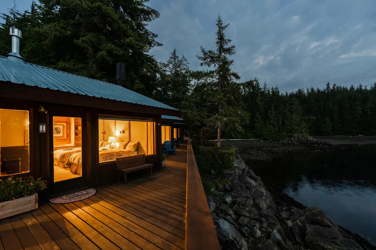 A guest room lit from within at dusk, seen from the cedar boardwalk that runs along the rocky shoreline