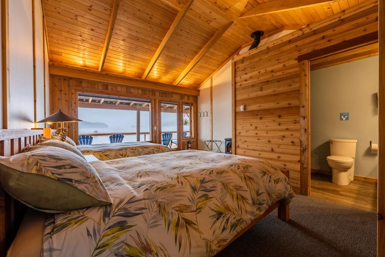 Guest room with two beds under a cedar ceiling, a wall of windows onto the harbour and the ensuite bathroom beyond