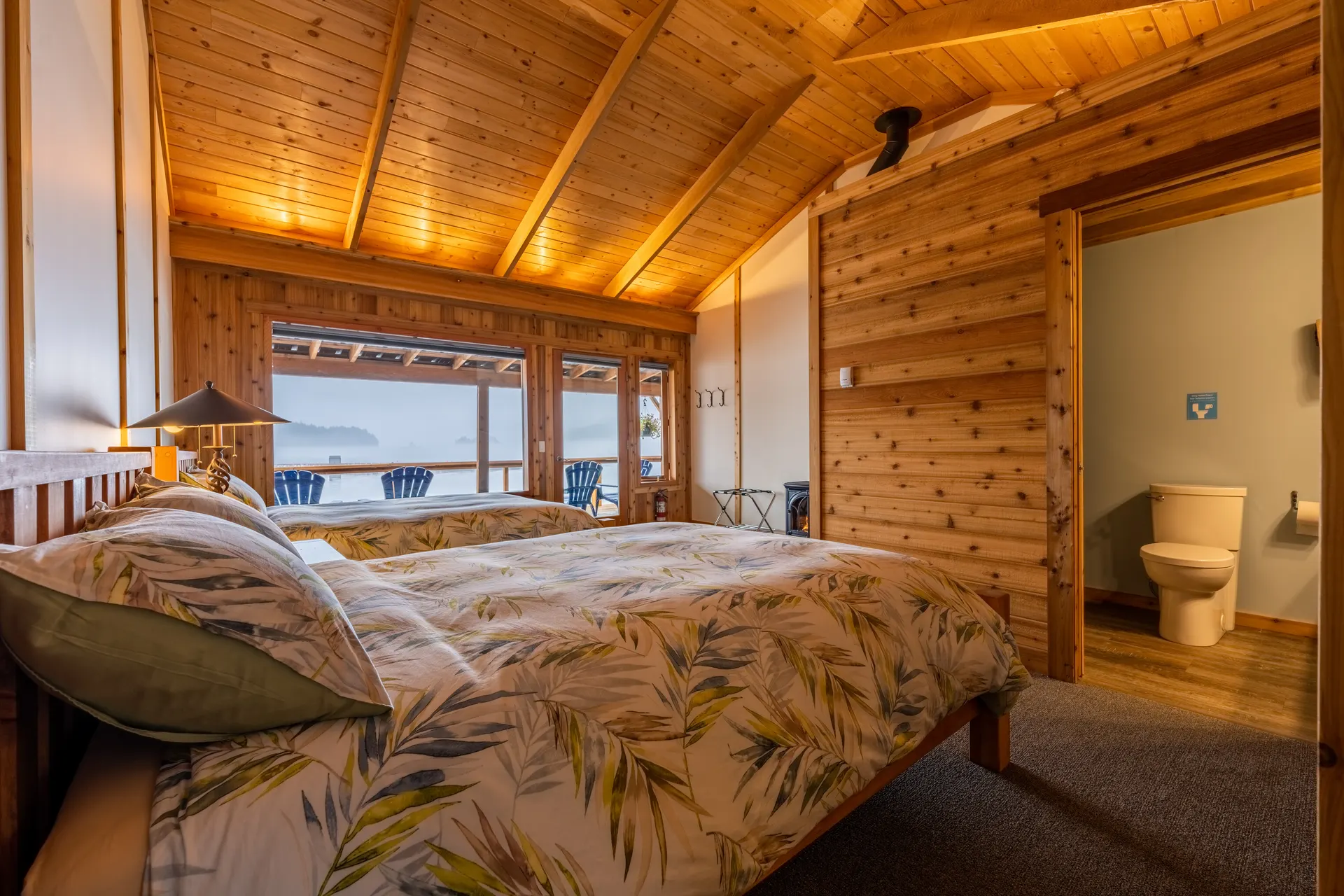 Guest room with two beds under a cedar ceiling, a wall of windows onto the harbour and the ensuite bathroom beyond