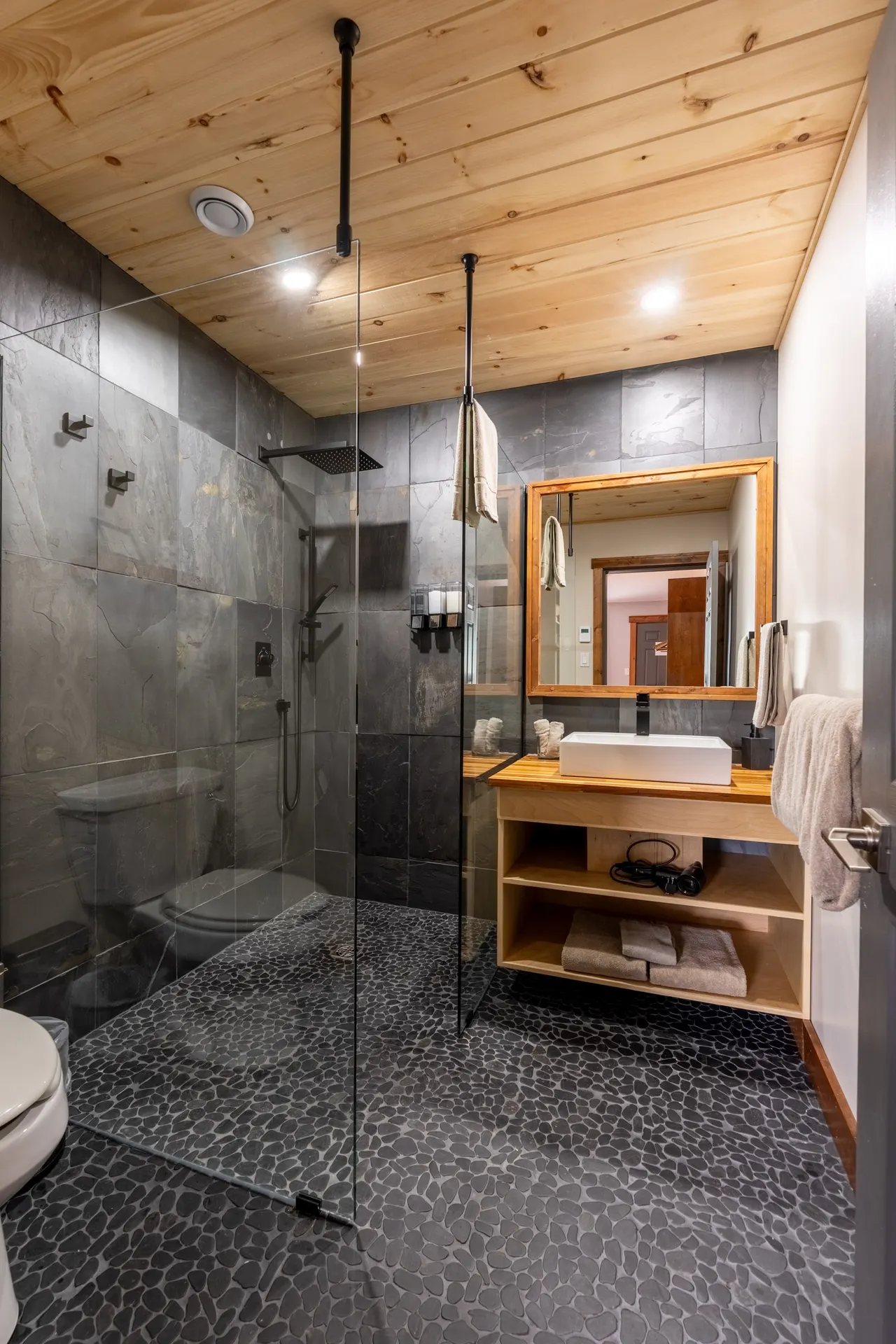 A guest bathroom at Knight Inlet Lodge: slate-tiled walk-in shower behind glass, a pebble floor, a pine ceiling and a live-edge vanity