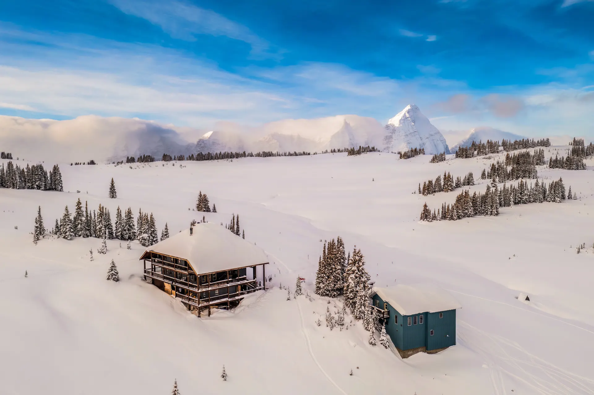 Purcell Mountain Lodge and its chalet from the air in deep winter, ski tracks on the bench and a wall of cloud-wrapped peaks behind under a dark blue sky