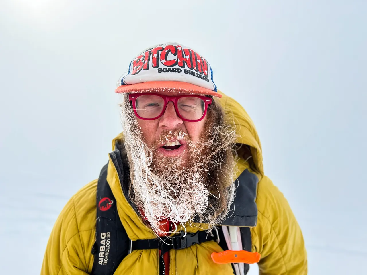 A ski tourer in a yellow jacket and an avalanche airbag pack, beard and eyelashes white with frost, grinning under a trucker cap on a whiteout day