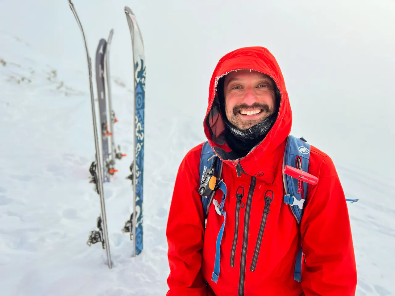 A skier in a red shell with the hood up, smiling on a snow slope with two pairs of touring skis planted upright behind him
