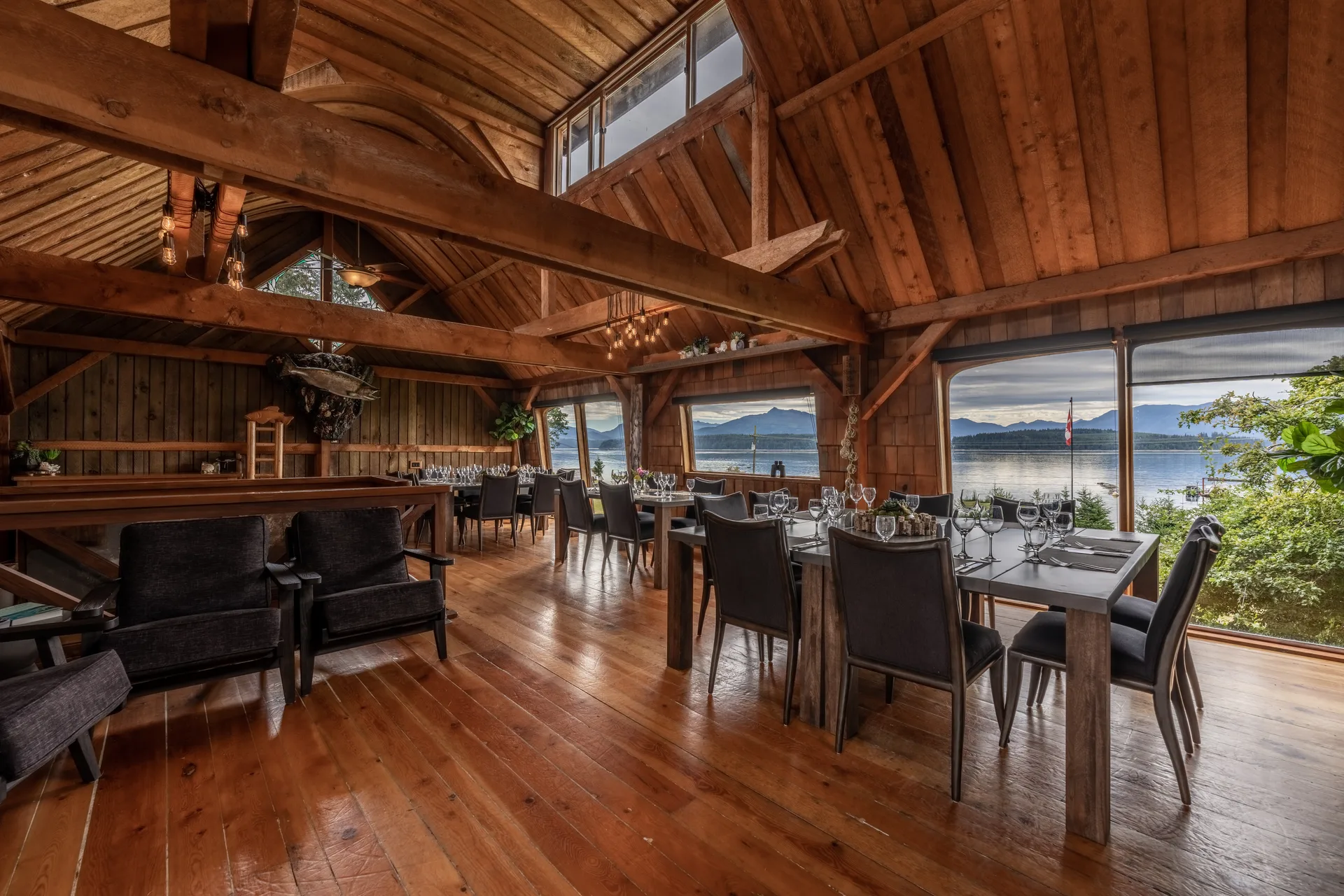 The lodge's timber great room with dining tables set for dinner and tall windows onto the water