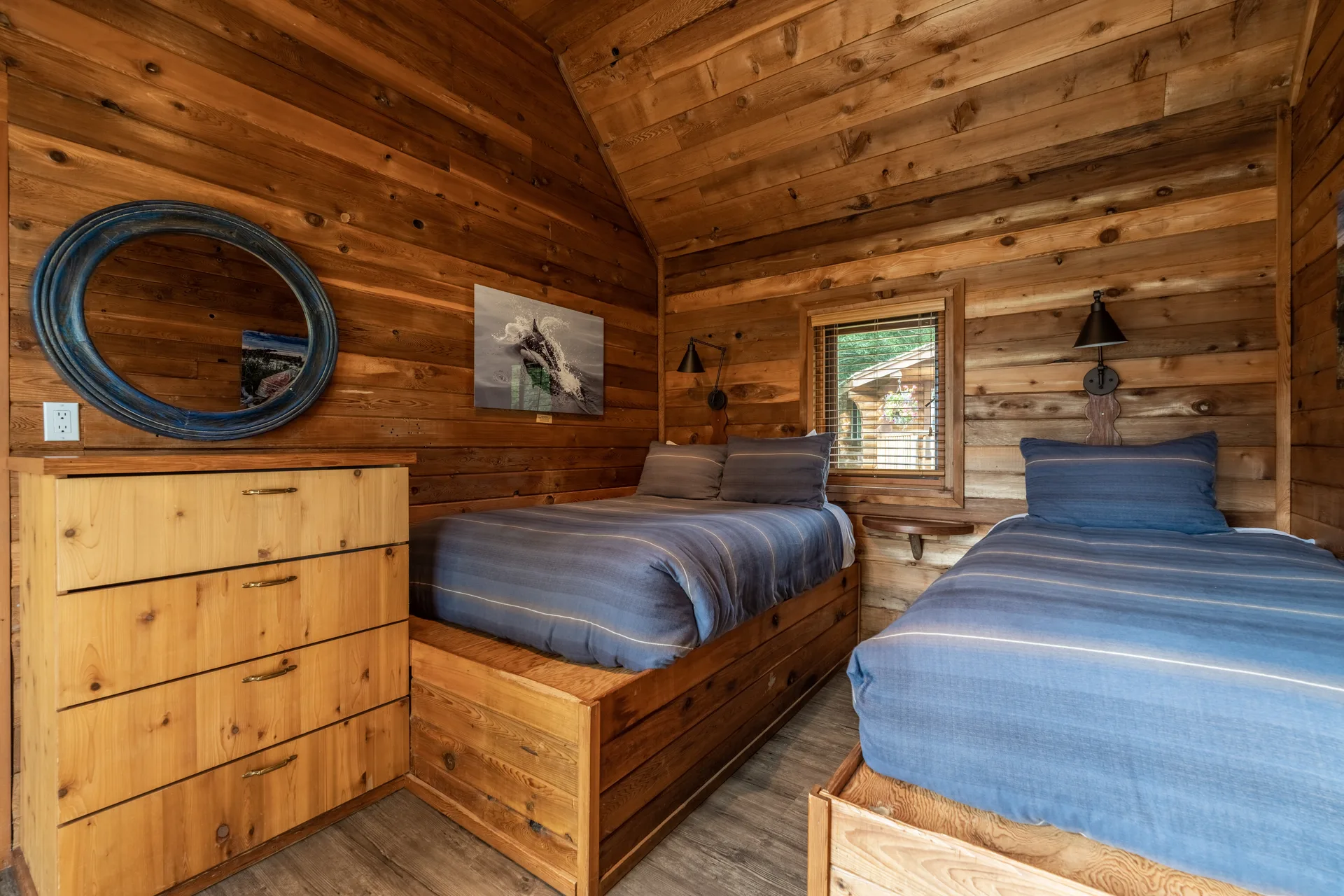 A log cabin bedroom with two twin beds in blue striped bedding beside a pine dresser