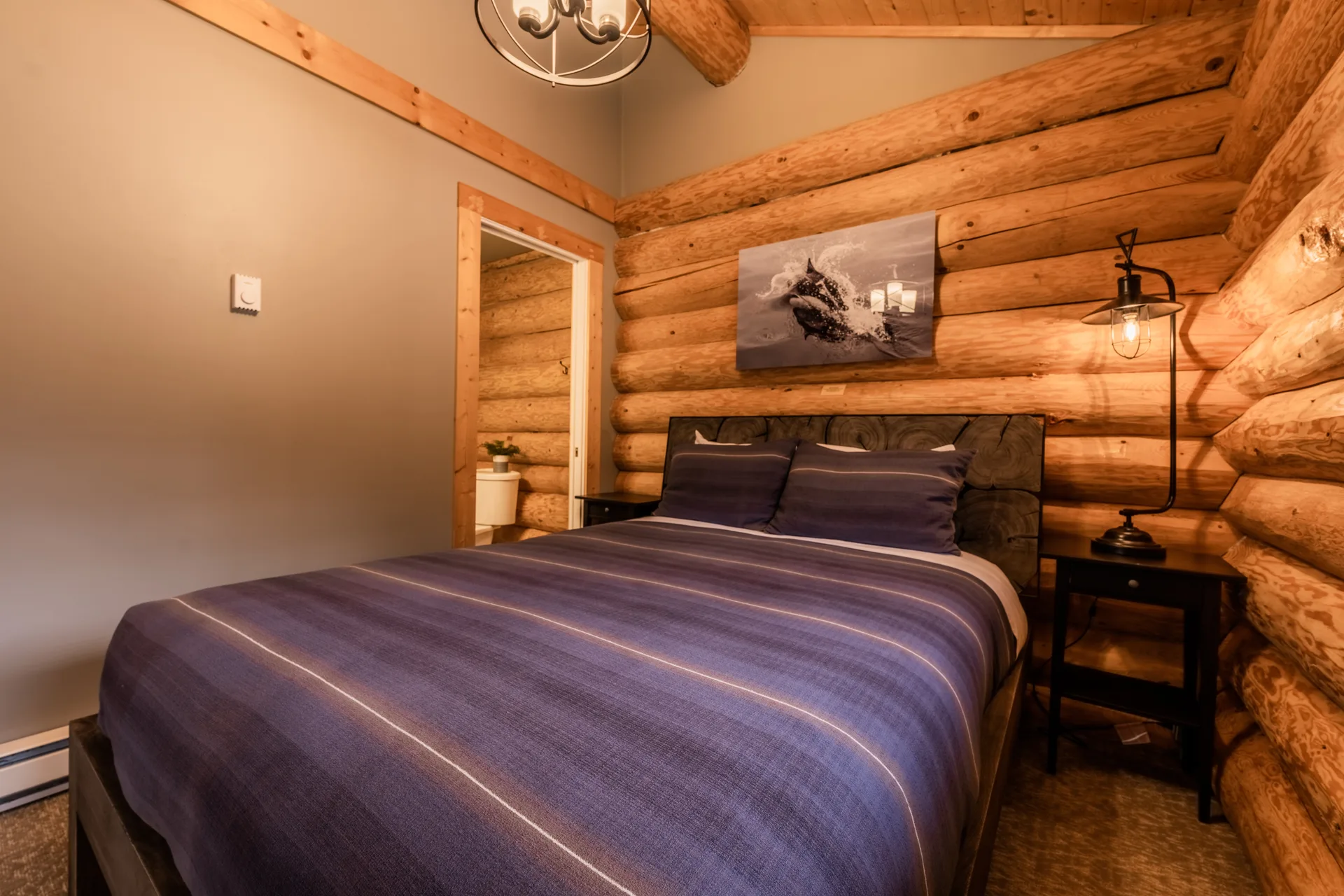Log cabin bedroom with a queen bed in purple bedding and a lit lamp on the night stand