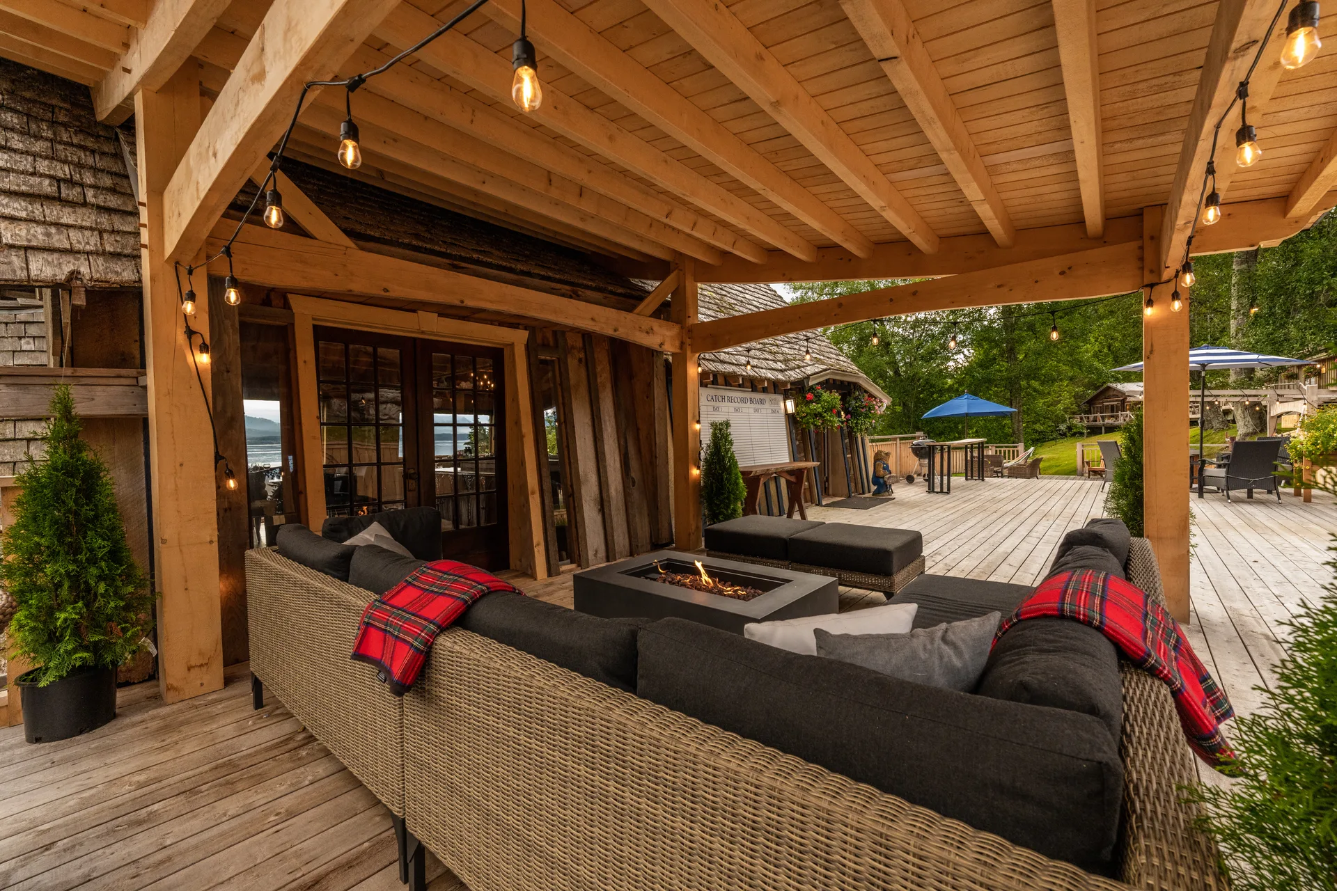 The covered deck at dusk with wicker sofas, a fire table and string lights, looking toward the lodge