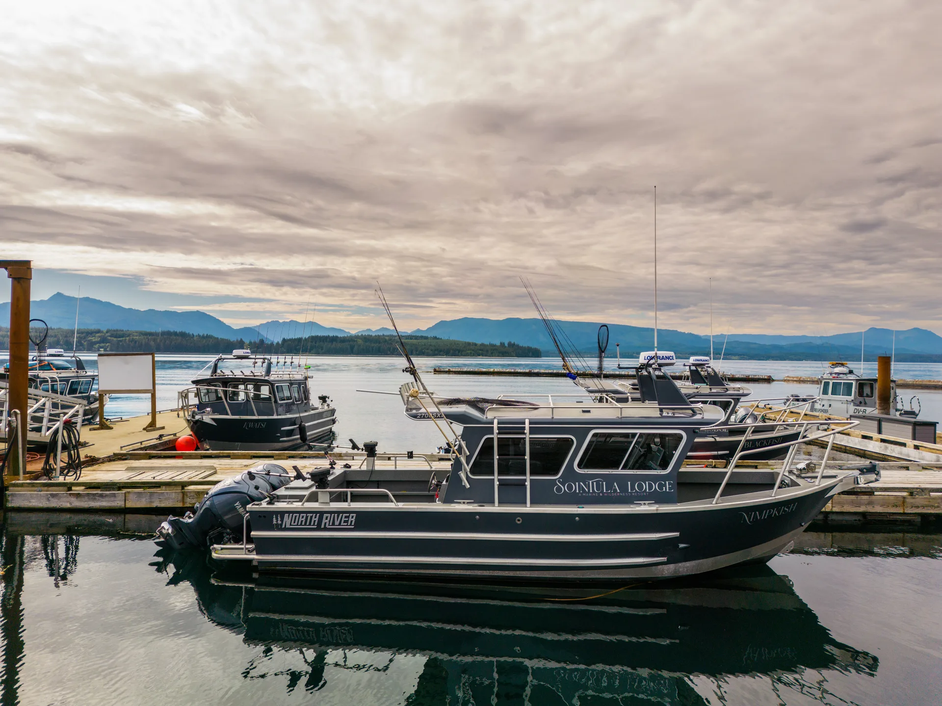 Two enclosed-cabin guide boats moored at the Sointula Lodge dock under a grey sky, with mountains across the strait