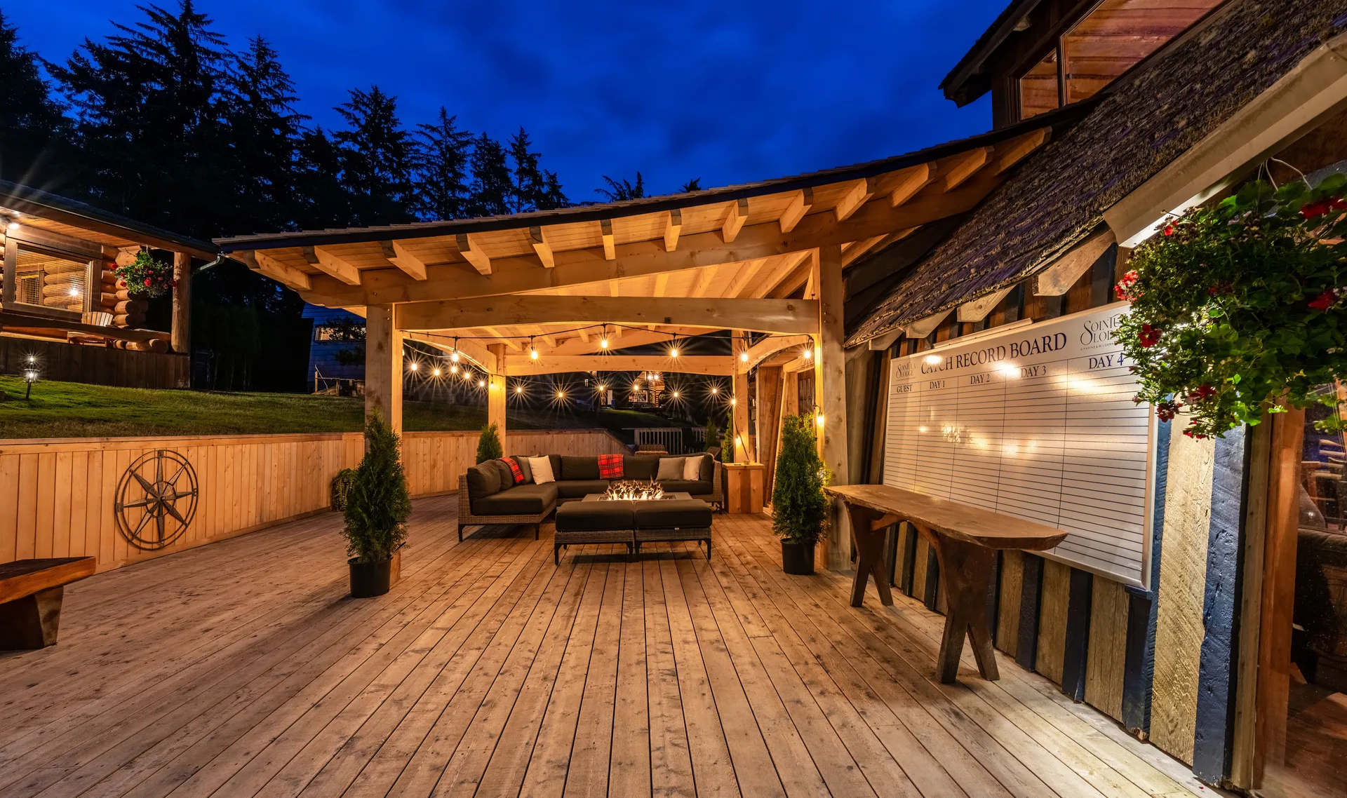 The lodge's covered timber deck lit with string lights at dusk, with sofas, a fire table and the lawn beyond
