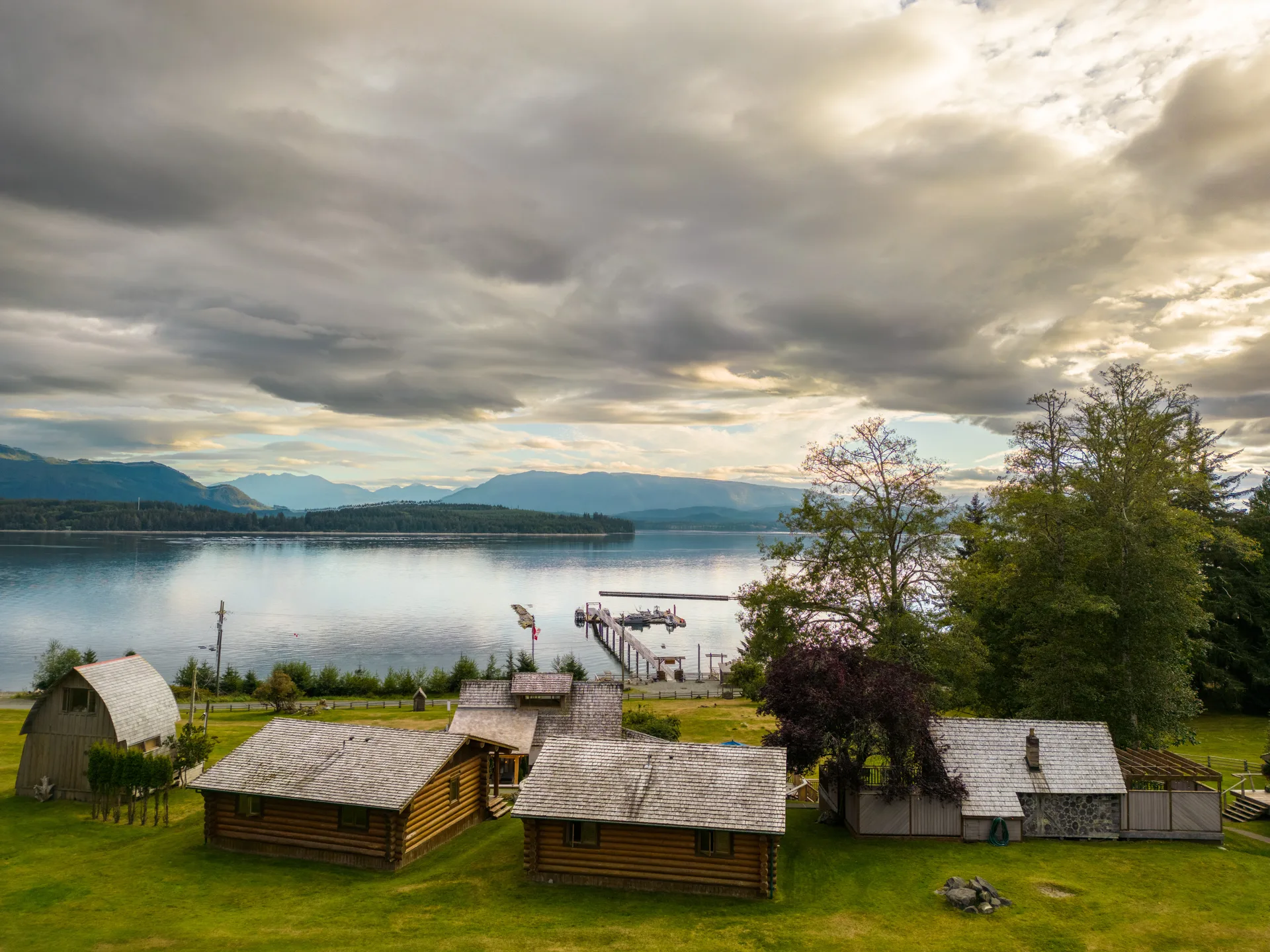 Aerial view of the log cabins and lawn with the dock and Broughton Strait beyond, under a dramatic evening sky