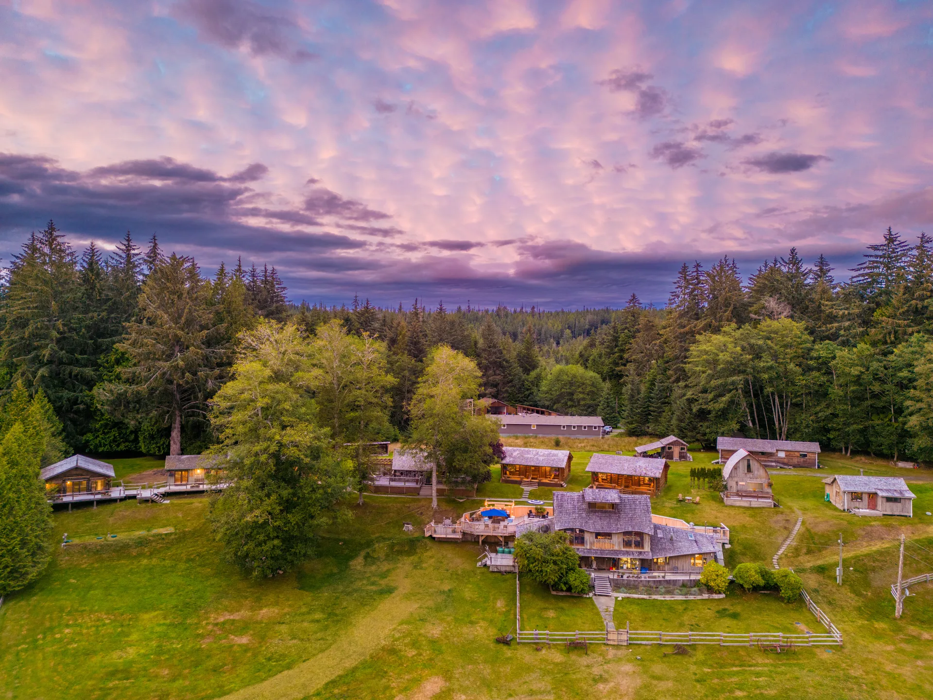 Aerial view of Sointula Lodge's cabins scattered across the lawn among tall conifers under a pink and purple dusk sky