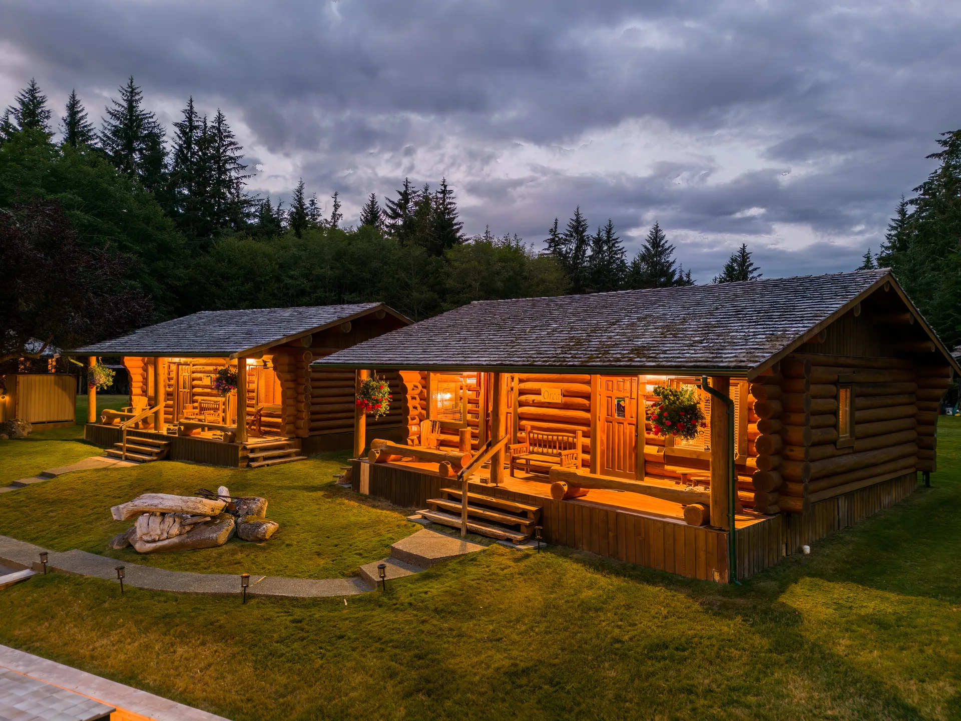Two log guest cabins with lit porches on the lawn at dusk, under a stormy sky