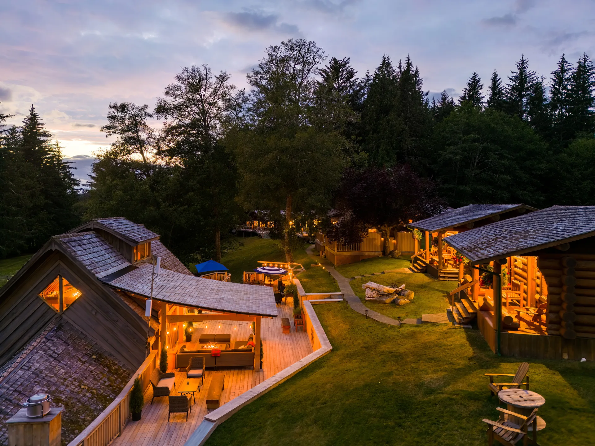 Aerial view at dusk of the lodge buildings, cabins and fire-pit patio glowing among the trees