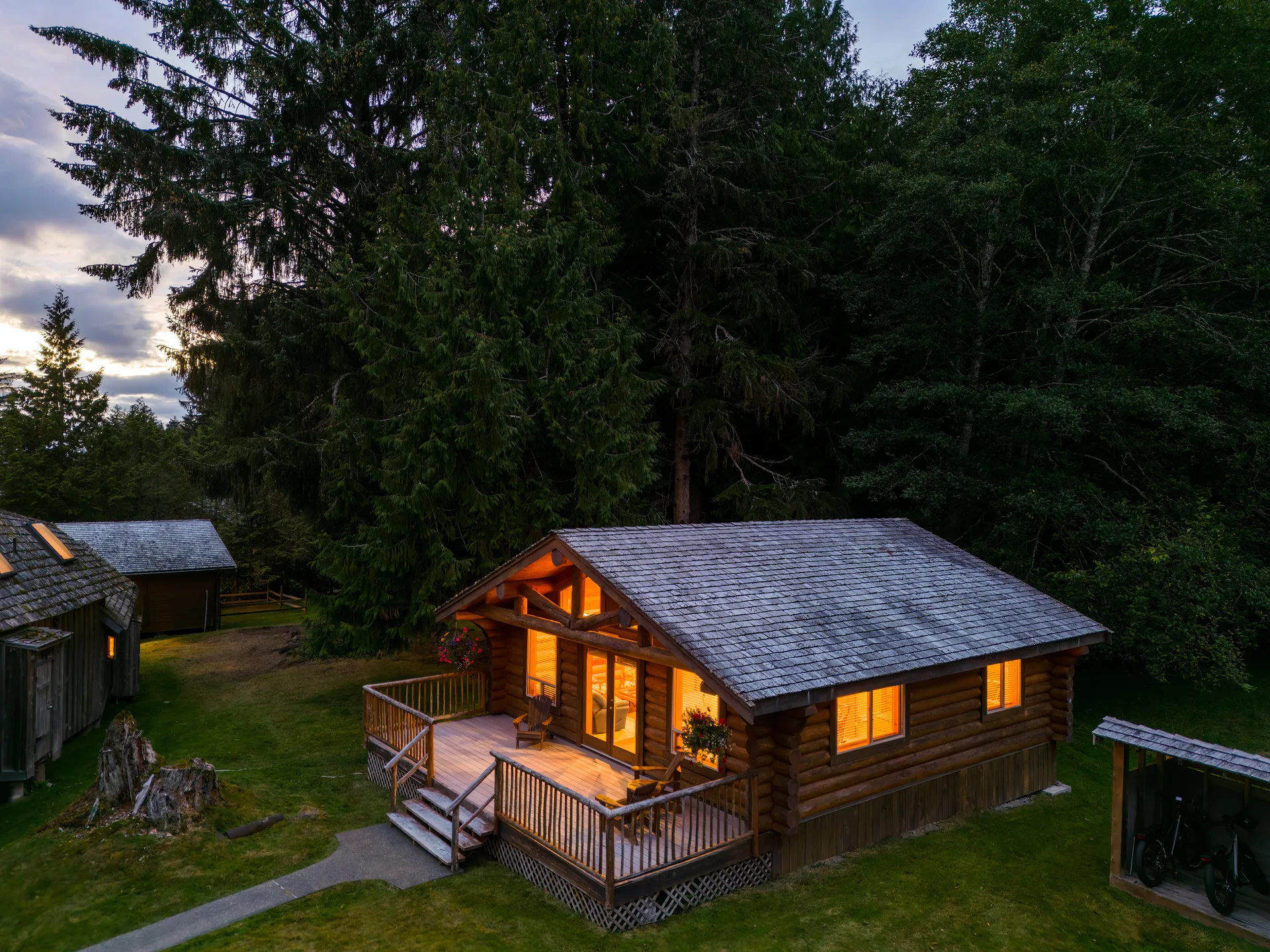 A log guest cabin with a lit porch and wooden steps at dusk, framed by tall cedars