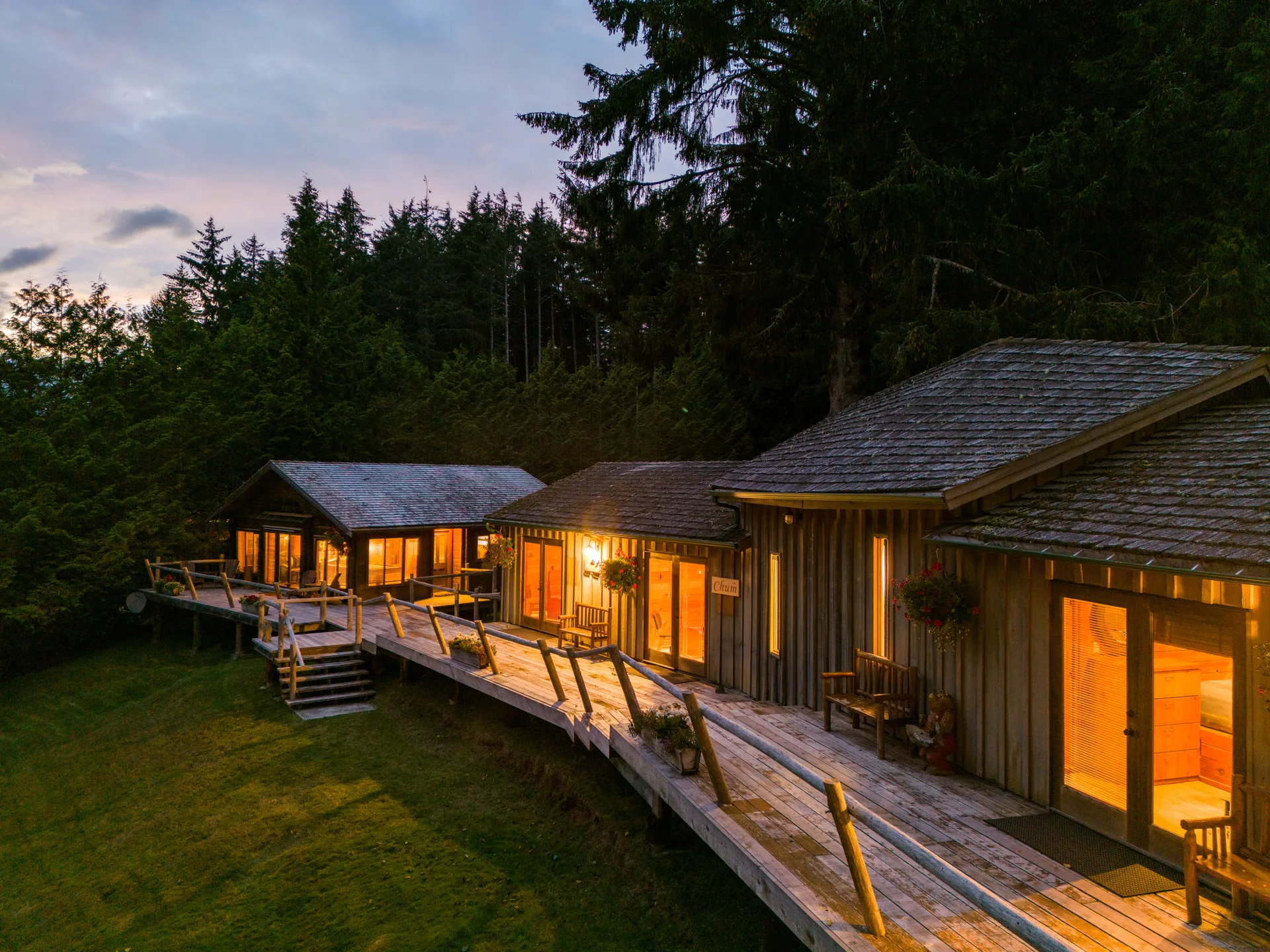 Two cabins with lit decks joined by a boardwalk at dusk, their glass doors glowing