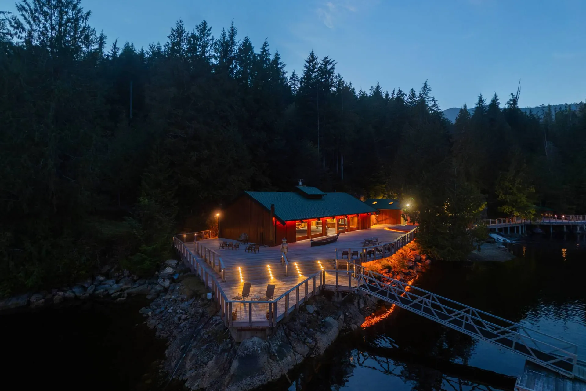 The longhouse and its deck lit at dusk above the water, the bridge to the floating lodge at the right