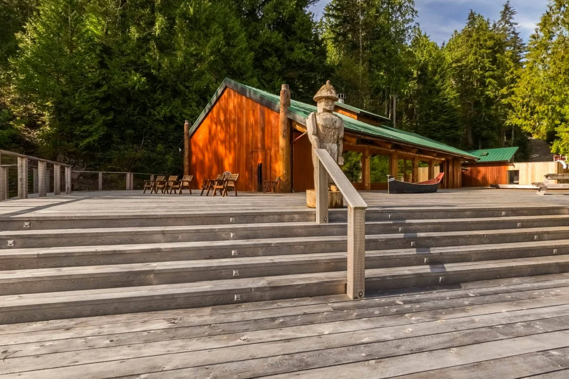 The deck of the longhouse in the sun, wide cedar steps, a carved post and chairs looking out over the bay