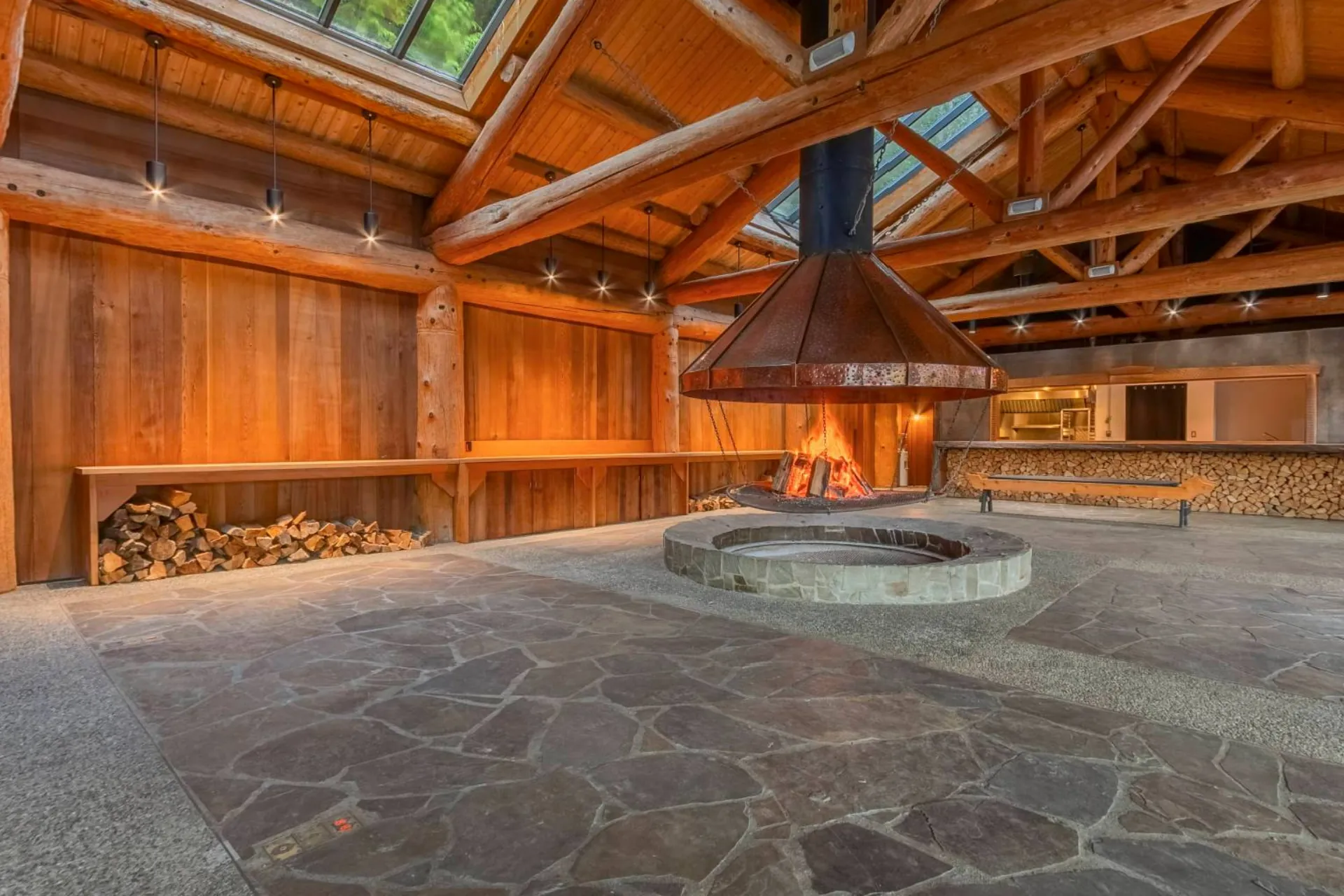 Inside the longhouse, the open fire pit under its copper hood, split wood stacked along the cedar wall