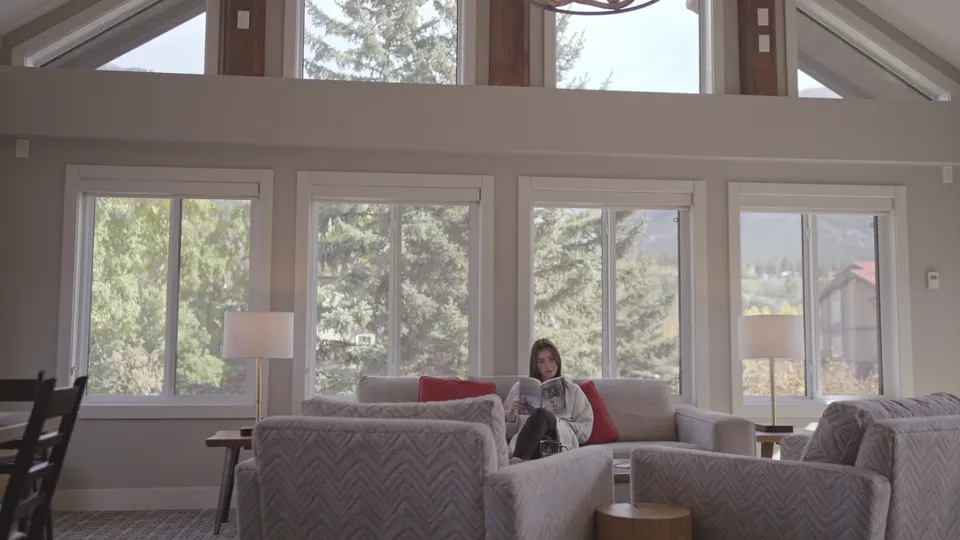 The great room under tall windows, armchairs and a dining table with the mountains outside