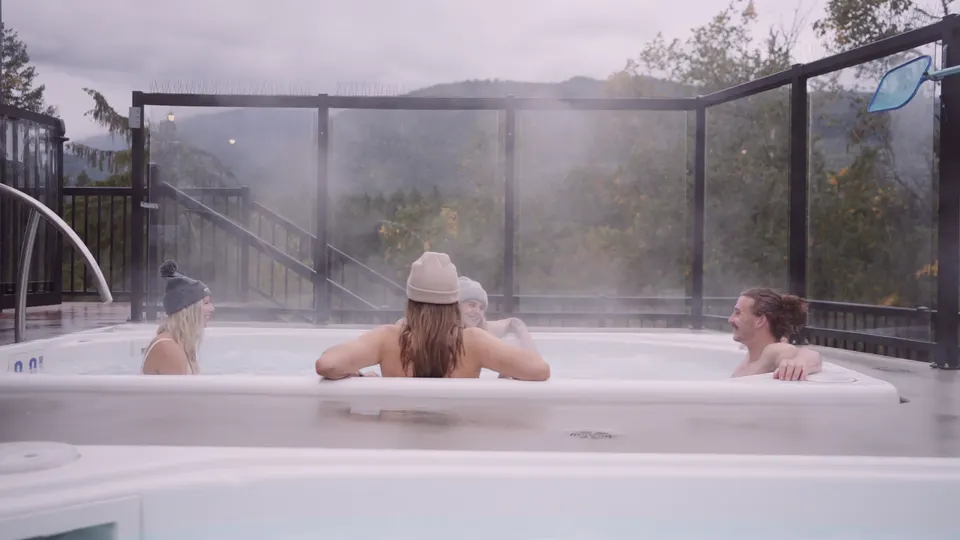 Three guests in the rooftop hot tub, steam rising, the glass rail and the mountains behind