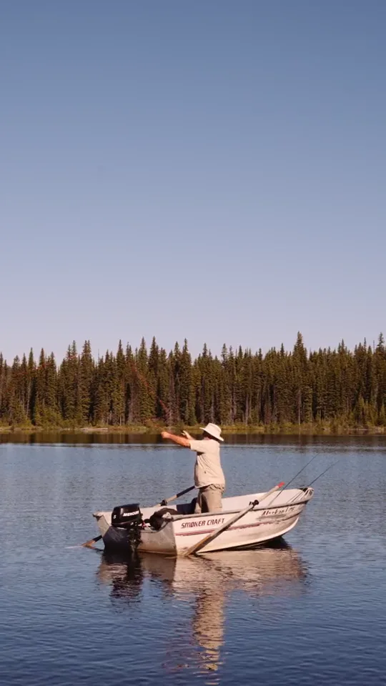 An angler casting from a small aluminium boat on a still lake, the forested shore across the water