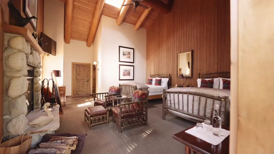 A log-walled guest room with a stone fireplace, a timber bed and a pair of armchairs in warm light