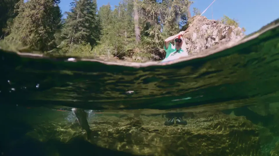 An angler casting from a granite boulder in a clear river, the camera half under the water