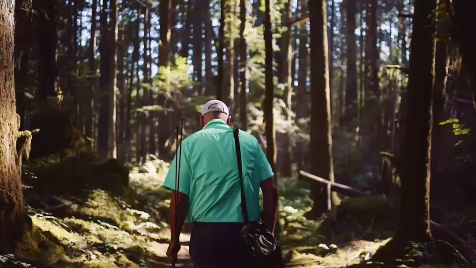 An angler in a green shirt walking a forest trail with a fly rod over his shoulder, sunlight through the cedars