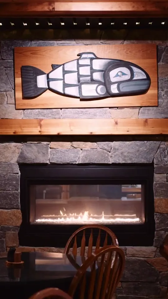 A carved salmon in First Nations formline style above the stone fireplace in the dining room, the gas fire lit below it