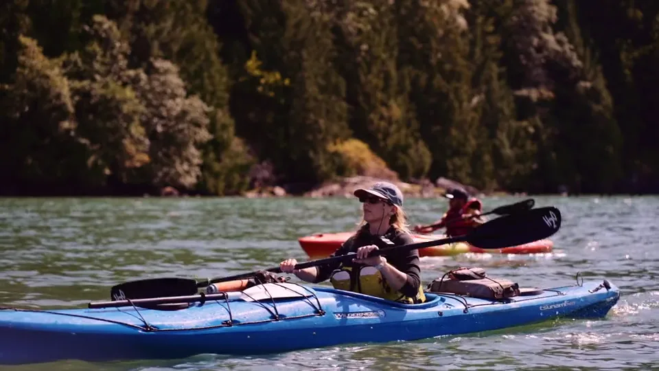 Two guests paddling a blue double kayak on the flat water of the cove, forest close behind them