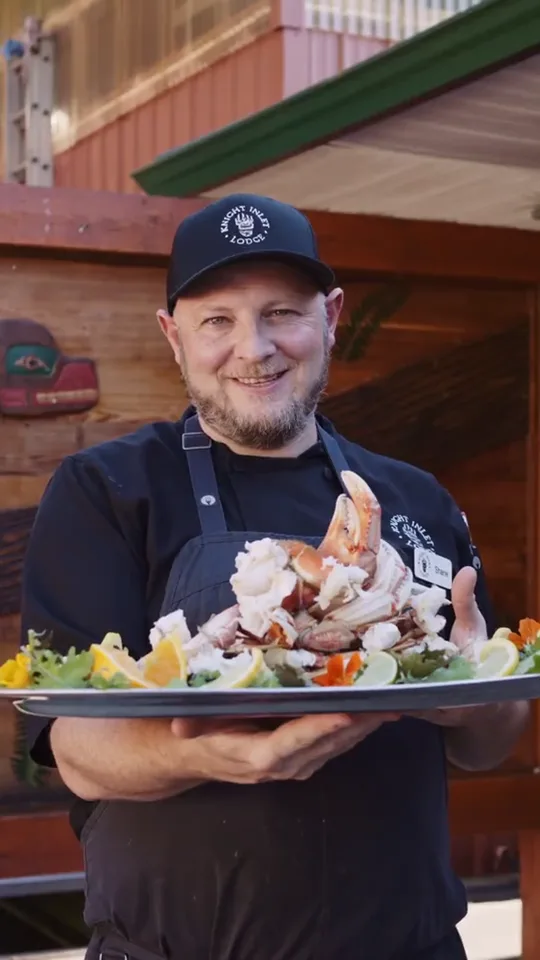 The chef in a black cap smiling as he carries a loaded platter out to the deck