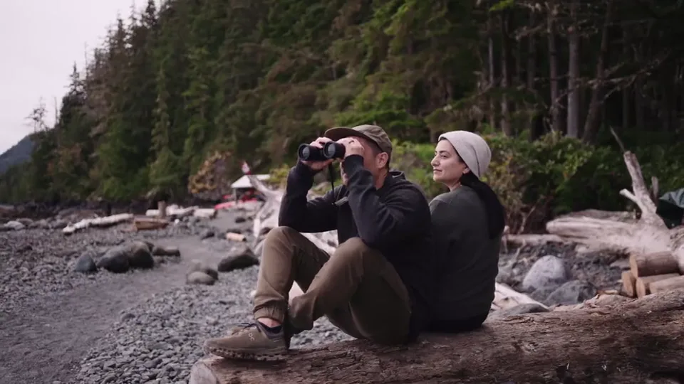 Two guests on a driftwood log on the pebble beach, one looking through binoculars