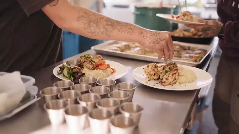 The camp chef reaching across plated dinners on the kitchen counter, a row of steel cups in front