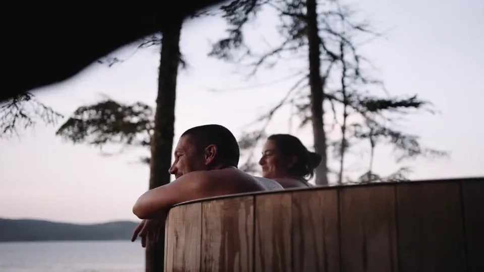 A couple in the wood-fired hot tub at sunset, the strait and the far shore behind them