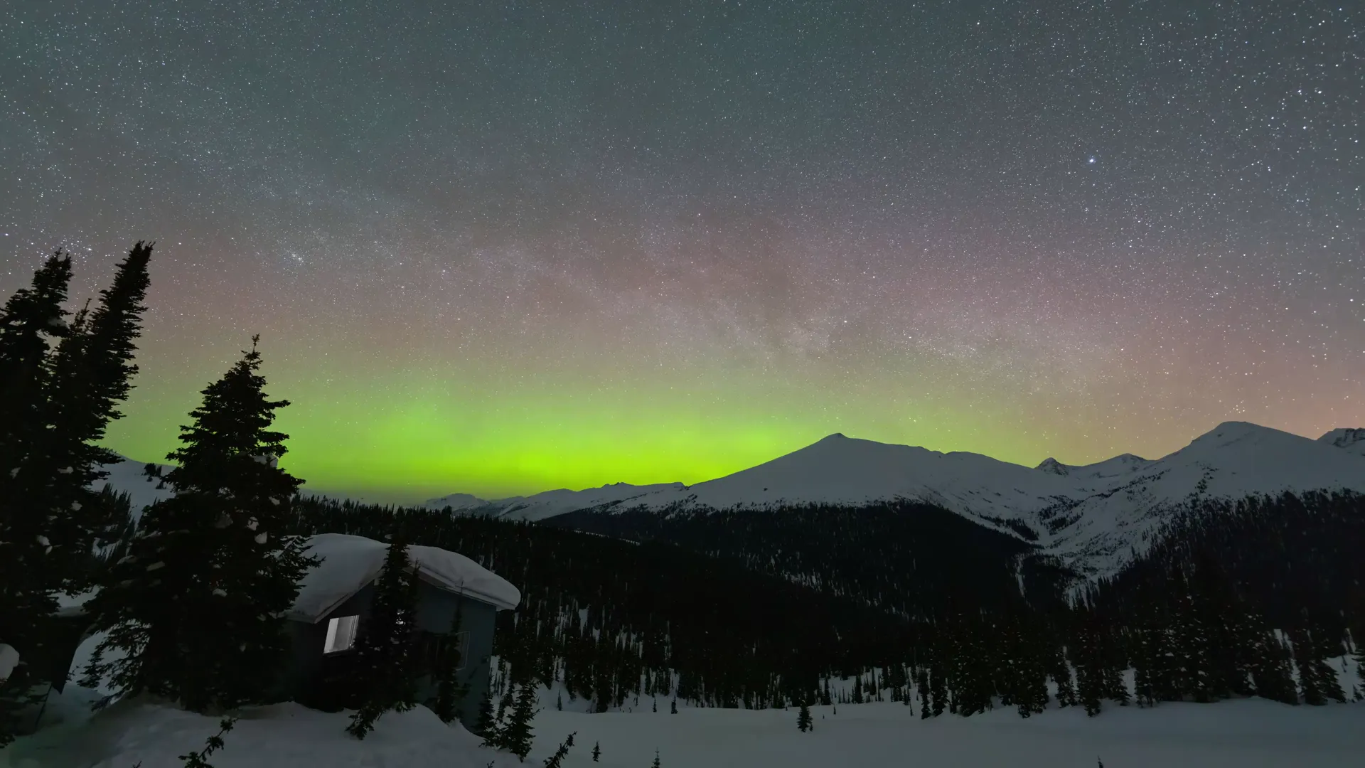 Green aurora and the Milky Way over Purcell Mountain Lodge at night, the lodge lit on its snow-covered bench