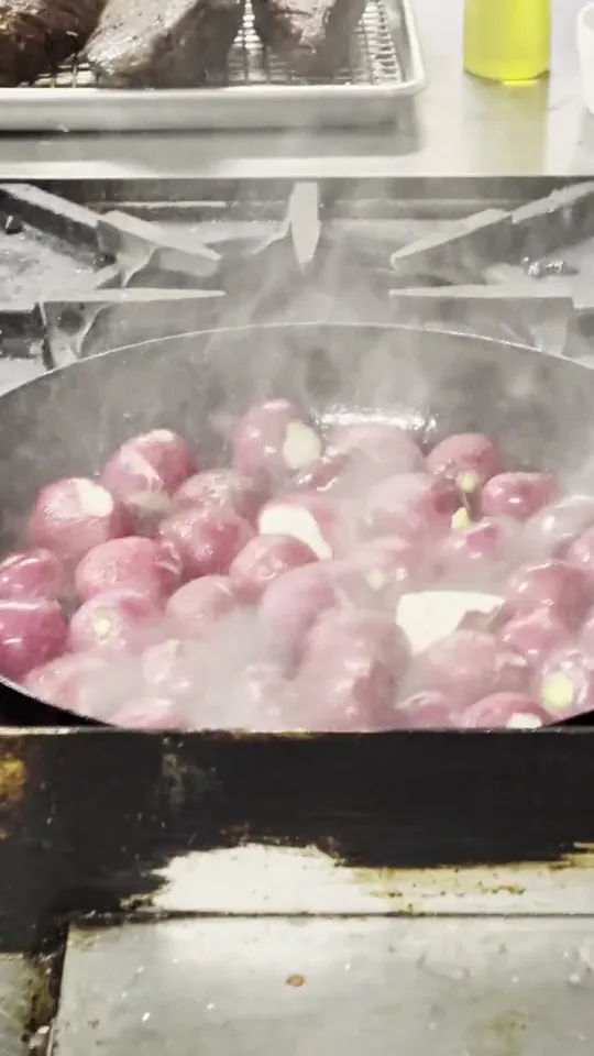 Flames and steam off a pan of purple onions on the lodge stove, a tray of seared meat behind
